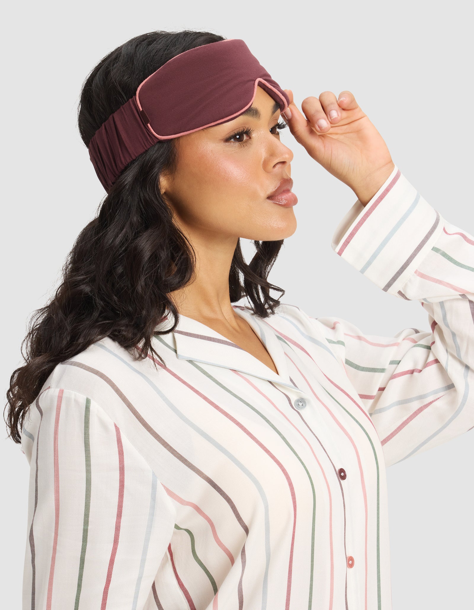 A woman in striped pajamas adjusts a Cozy Earth Bamboo Stretch-Knit Sleep Mask on her forehead, gazing to the side against a plain light background.
