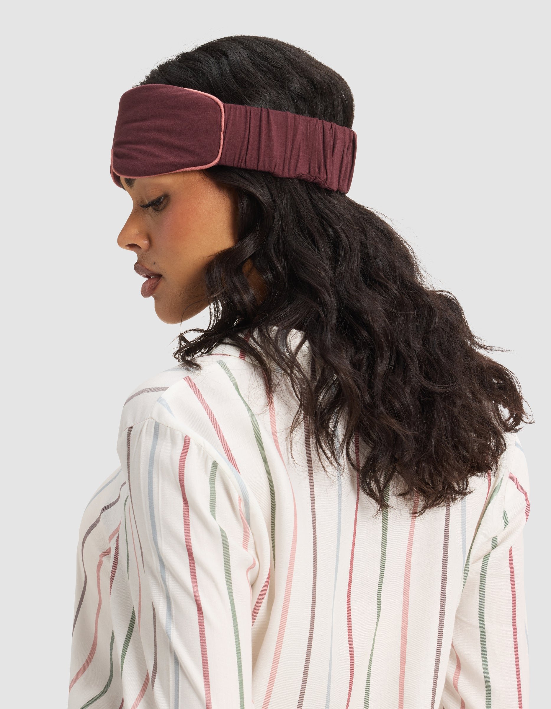Woman with wavy dark hair wears the Cozy Earth Bamboo Stretch-Knit Sleep Mask and a white shirt with pink and green stripes, facing sideways against a plain light background.