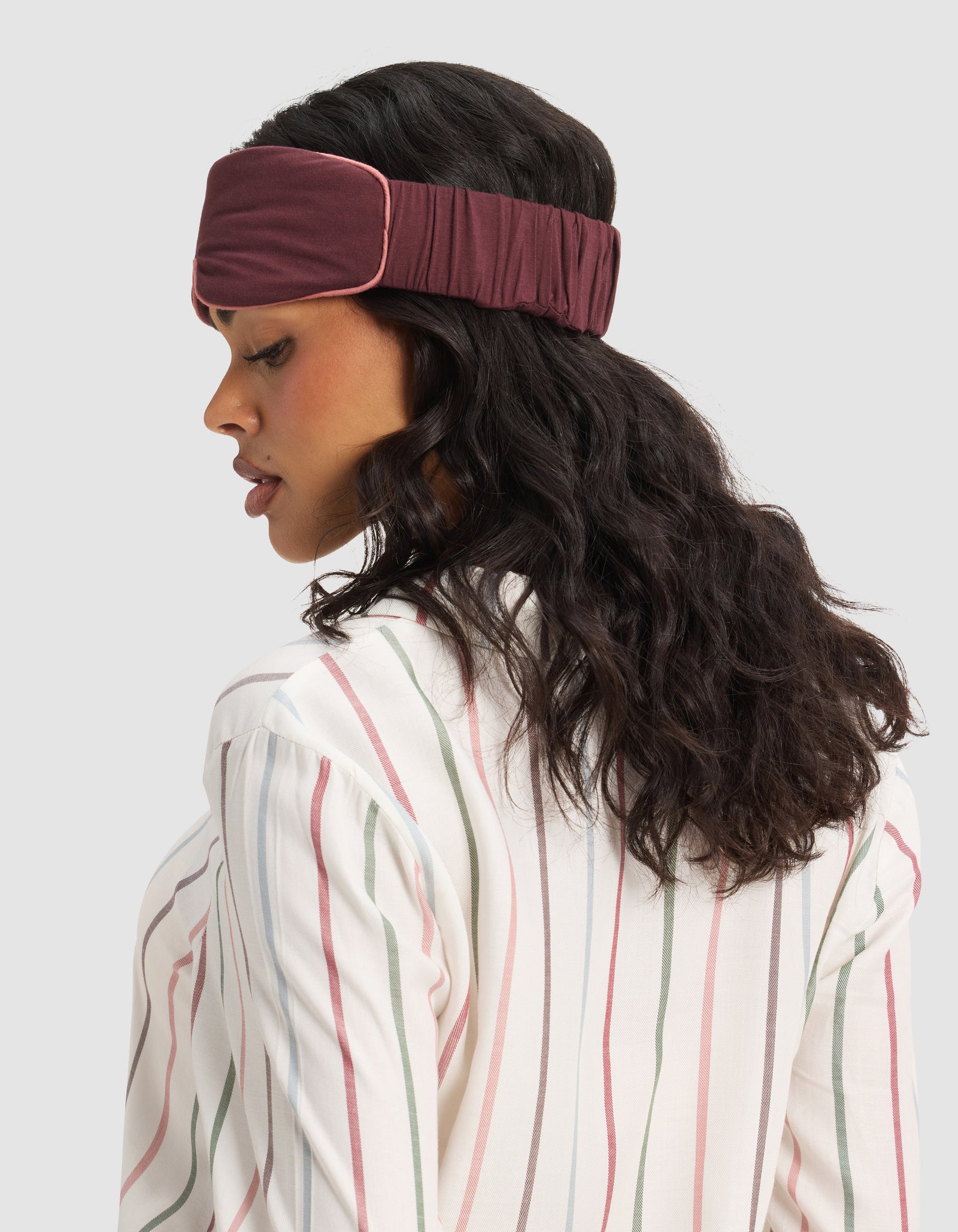 Woman with wavy dark hair wears the Cozy Earth Bamboo Stretch-Knit Sleep Mask and a white shirt with pink and green stripes, facing sideways against a plain light background.