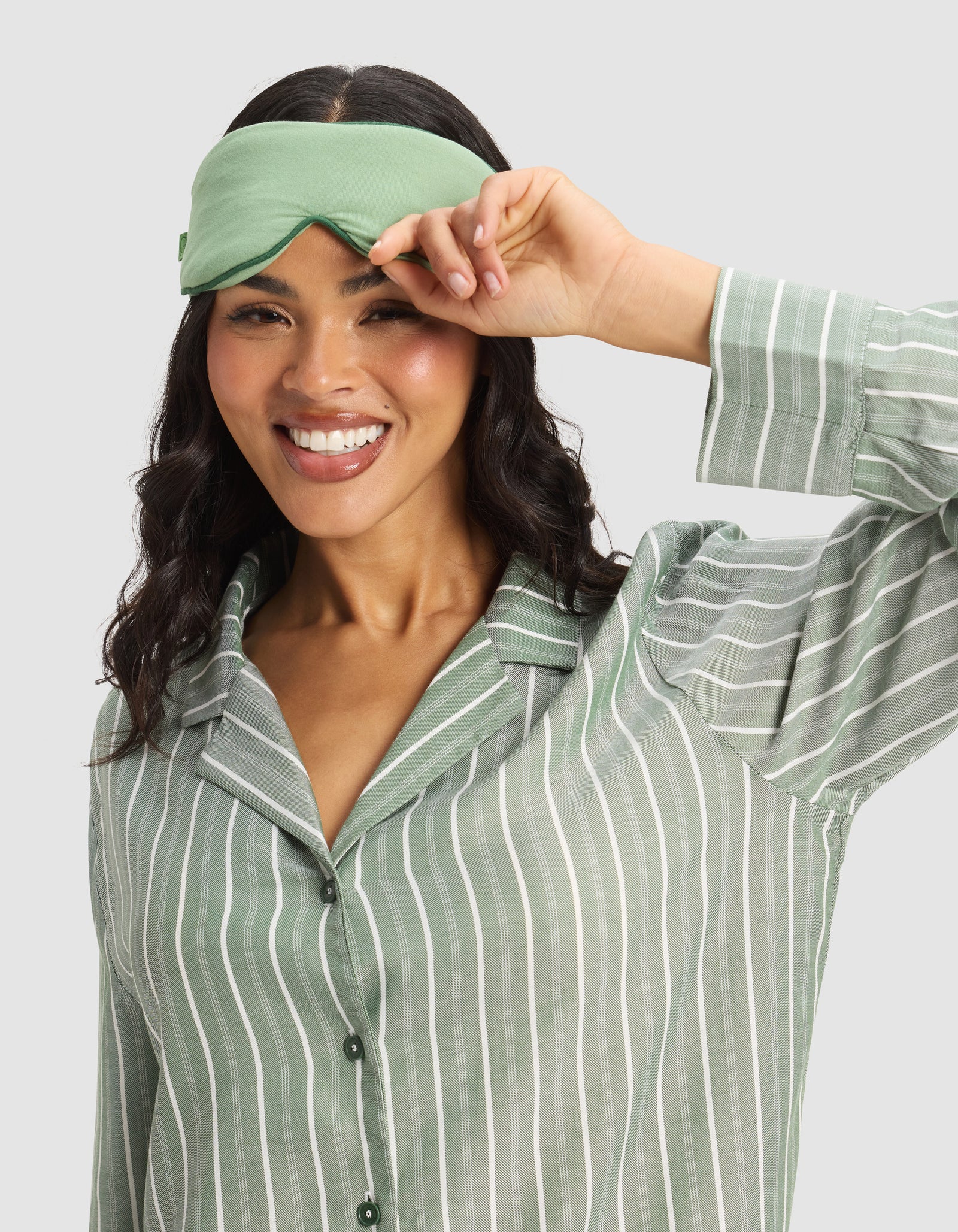 A woman smiles while lifting a Cozy Earth Bamboo Stretch-Knit Sleep Mask above her eyes, wearing a matching green and white striped pajama top.