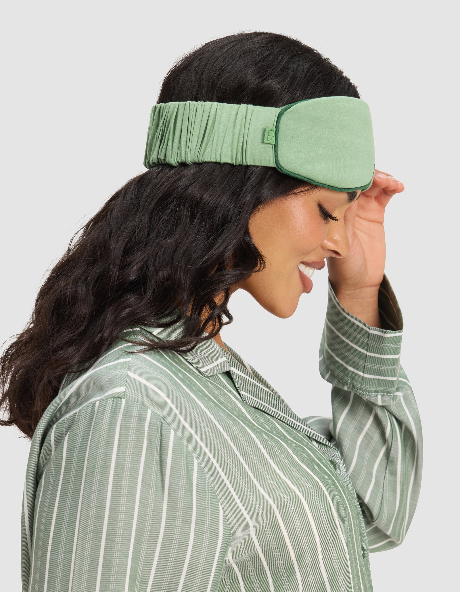 A woman with long dark hair smiles and touches a Cozy Earth Bamboo Stretch-Knit Sleep Mask while wearing a matching green striped pajama top. The background is light gray.