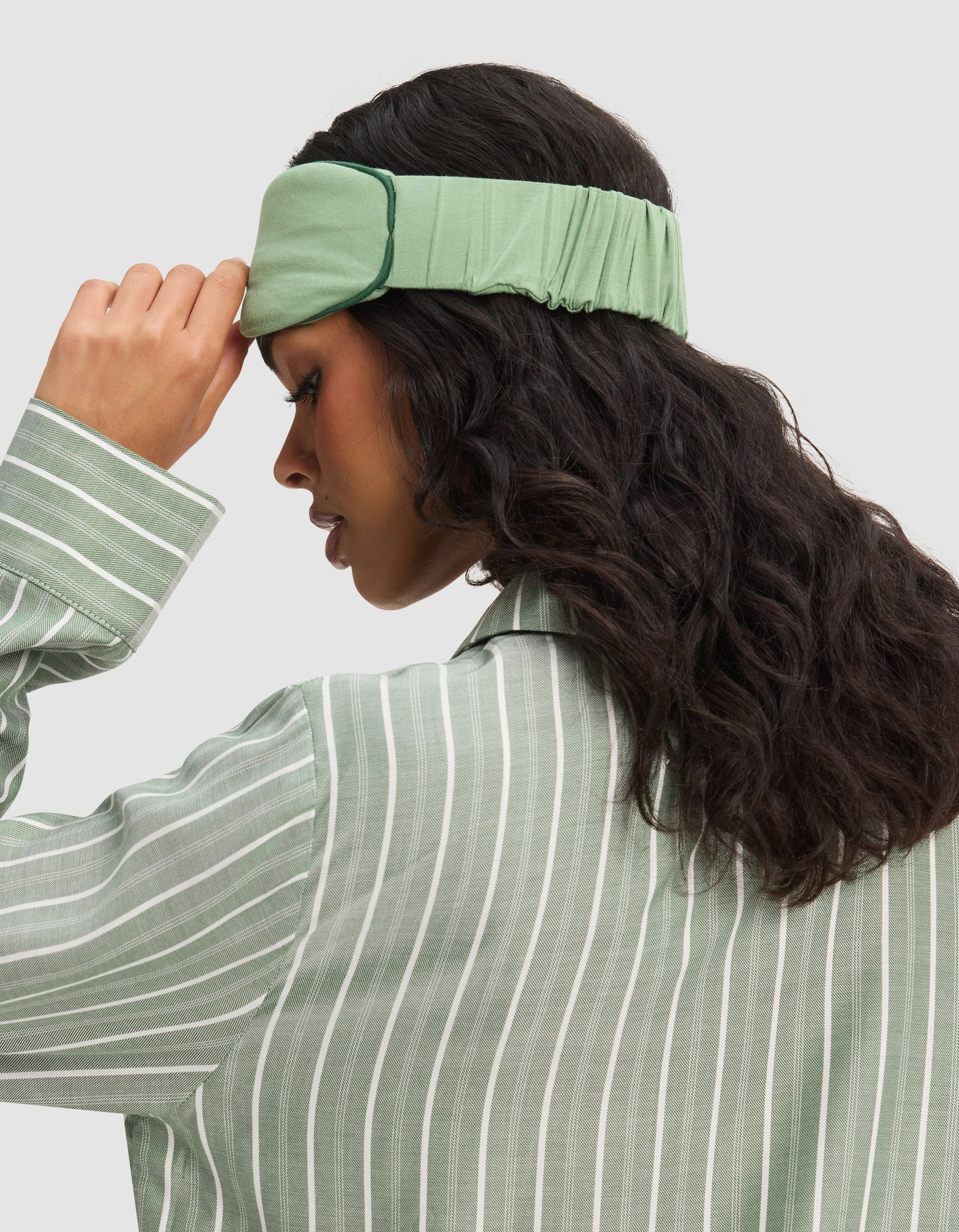 A woman with long, wavy dark hair adjusts a Cozy Earth Bamboo Stretch-Knit Sleep Mask while wearing a green striped pajama top. Her profile stands out against a plain background.