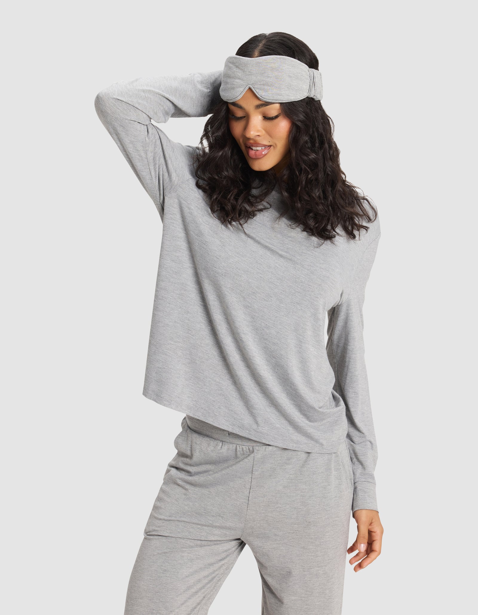 A woman poses with one hand on her head, smiling and looking down while wearing a Cozy Earth Bamboo Stretch-Knit Sleep Mask and a grey long-sleeve pajama set.