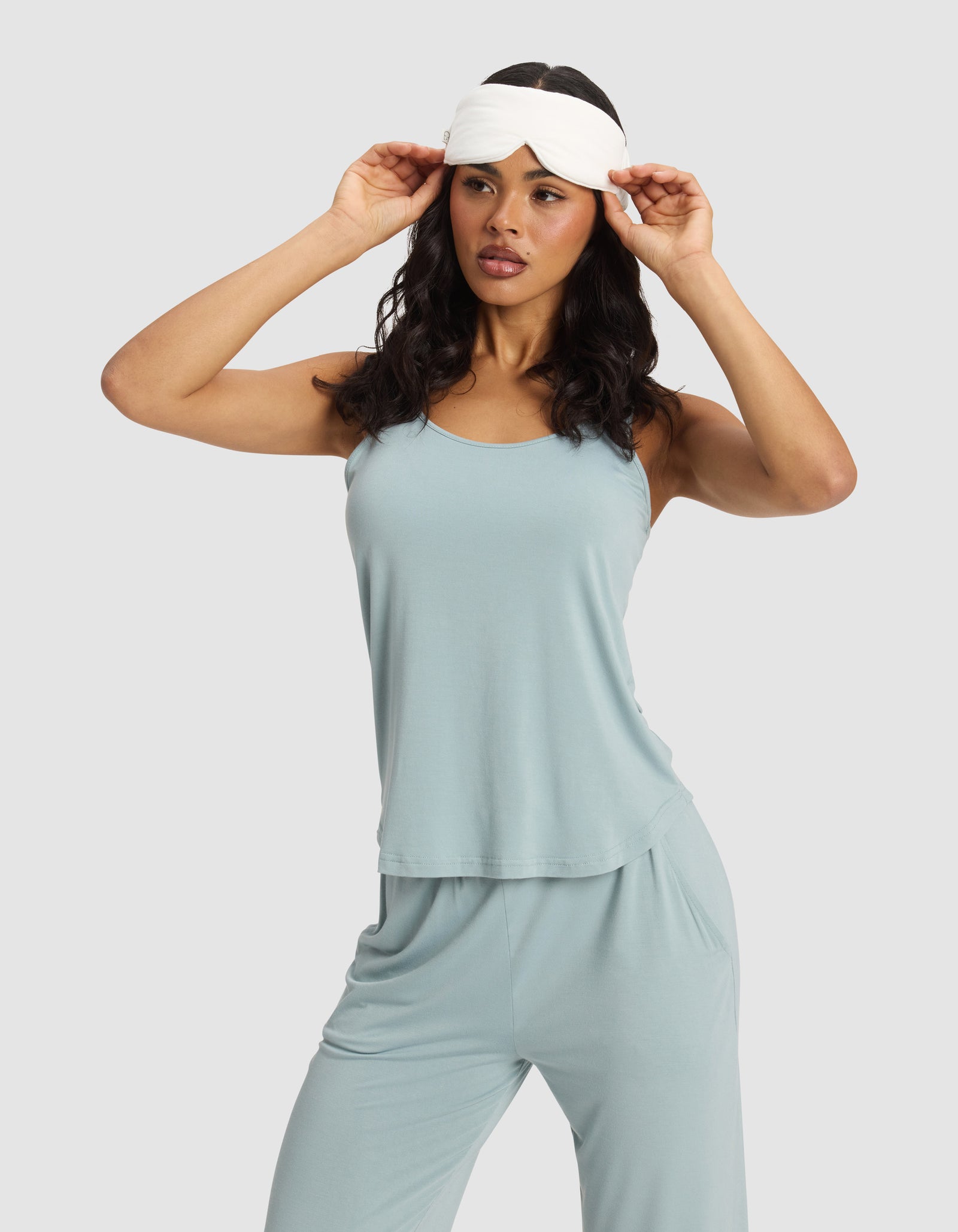 A woman in a light blue sleeveless pajama set adjusts a Cozy Earth Bamboo Stretch-Knit Sleep Mask on her forehead, standing against a plain background.