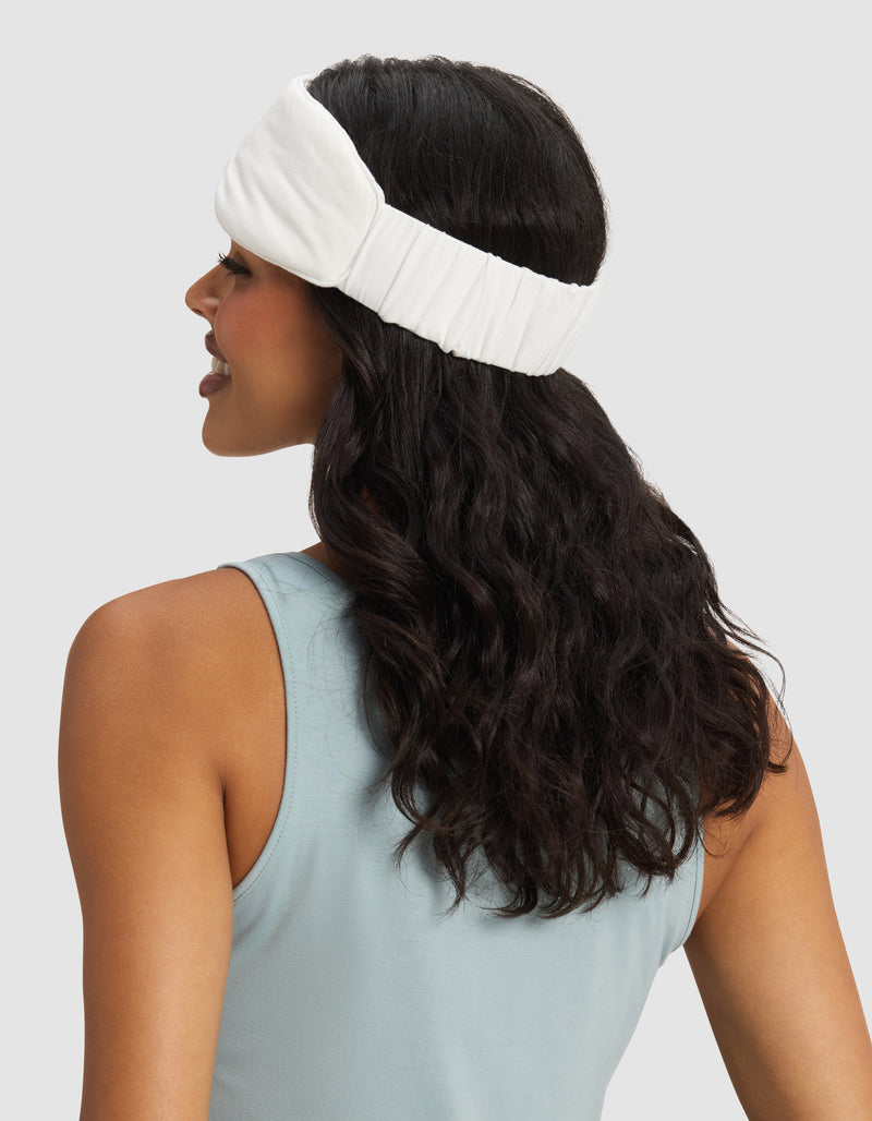 A woman with wavy dark hair faces away, wearing Cozy Earth's Bamboo Stretch-Knit Sleep Mask against a plain light background.