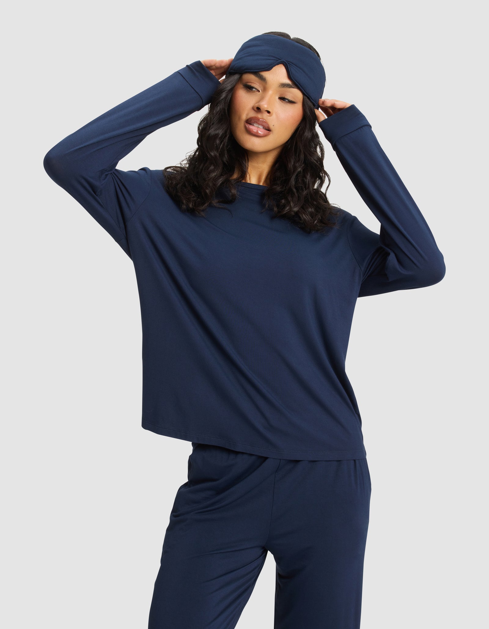A woman wearing a navy blue pajama set poses with her hands touching the sides of a Cozy Earth Bamboo Stretch-Knit Sleep Mask, standing against a plain light background.