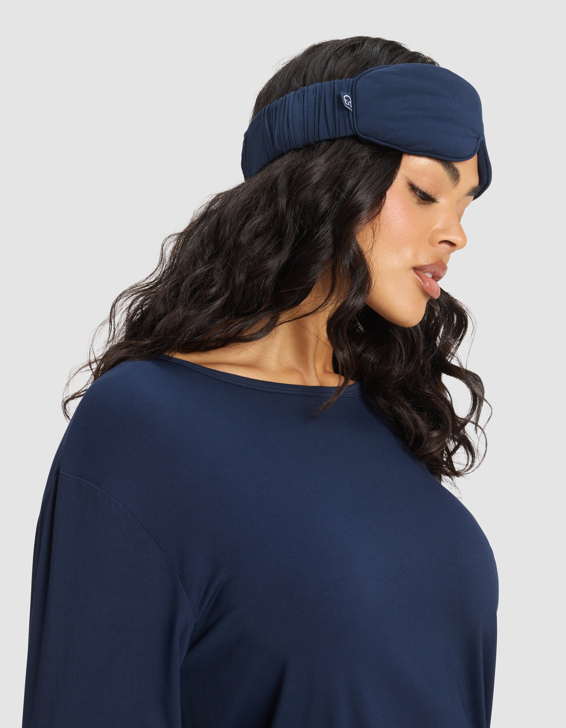 Woman with long wavy hair wearing a Cozy Earth Bamboo Stretch-Knit Sleep Mask and matching navy blue top, looking down to her left against a light gray background. 