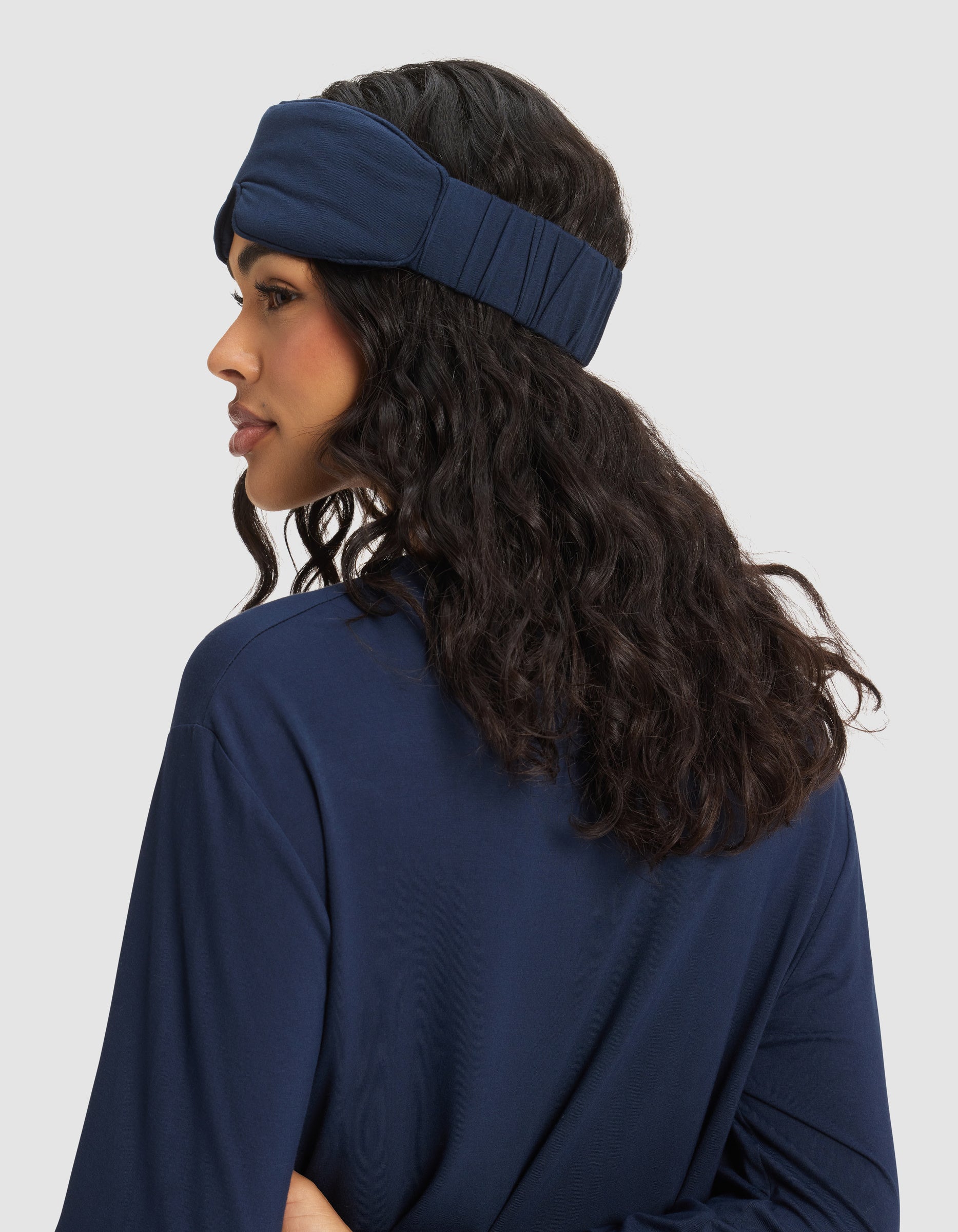 A woman with long, wavy dark hair is shown from the side and back wearing the Cozy Earth Bamboo Stretch-Knit Sleep Mask in navy blue, paired with a matching top against a plain light gray background.|Color:Navy