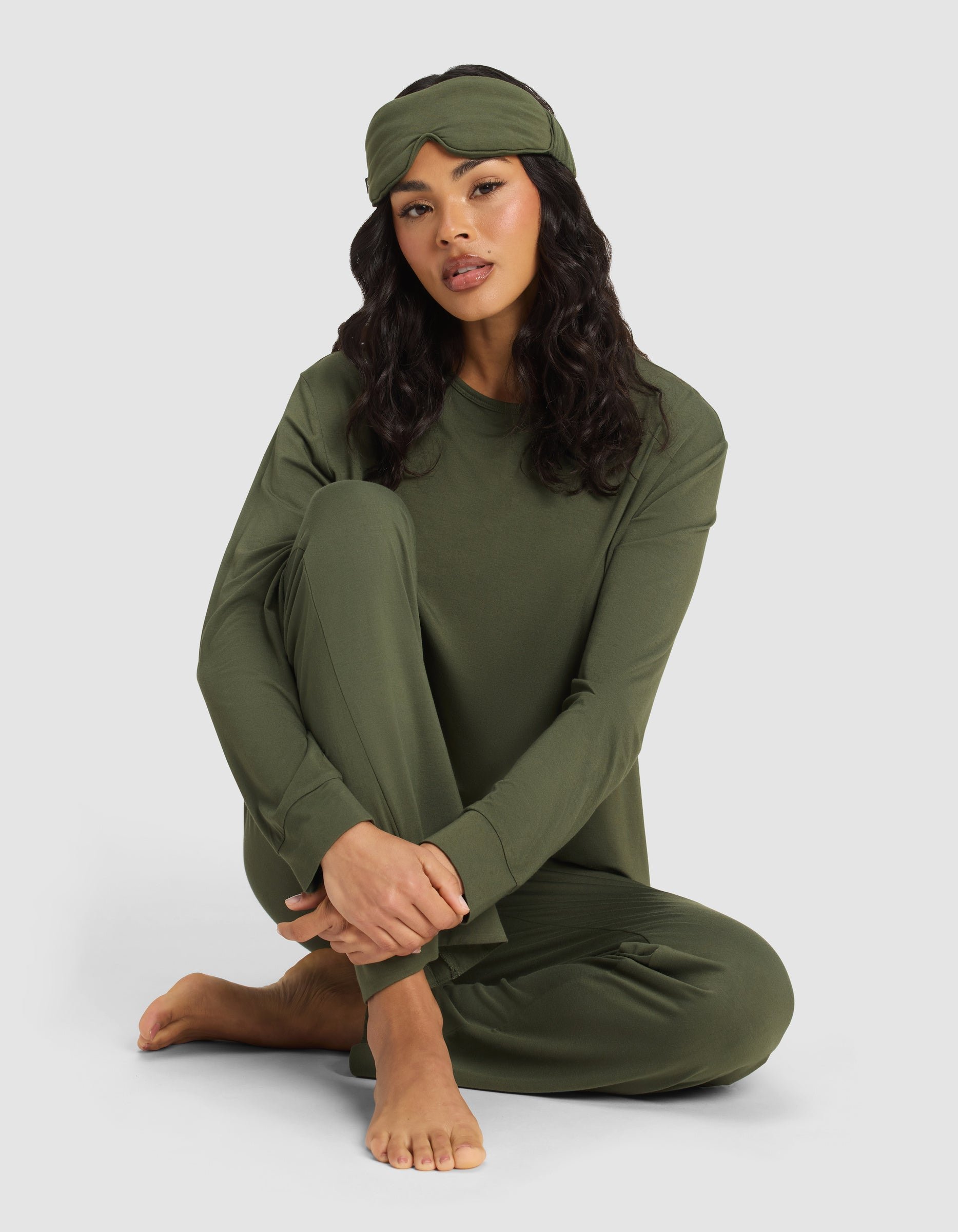 A woman with long dark hair sits barefoot on the floor, relaxed against a plain light background, wearing an olive green pajama set and the Cozy Earth Bamboo Stretch-Knit Sleep Mask on her head.