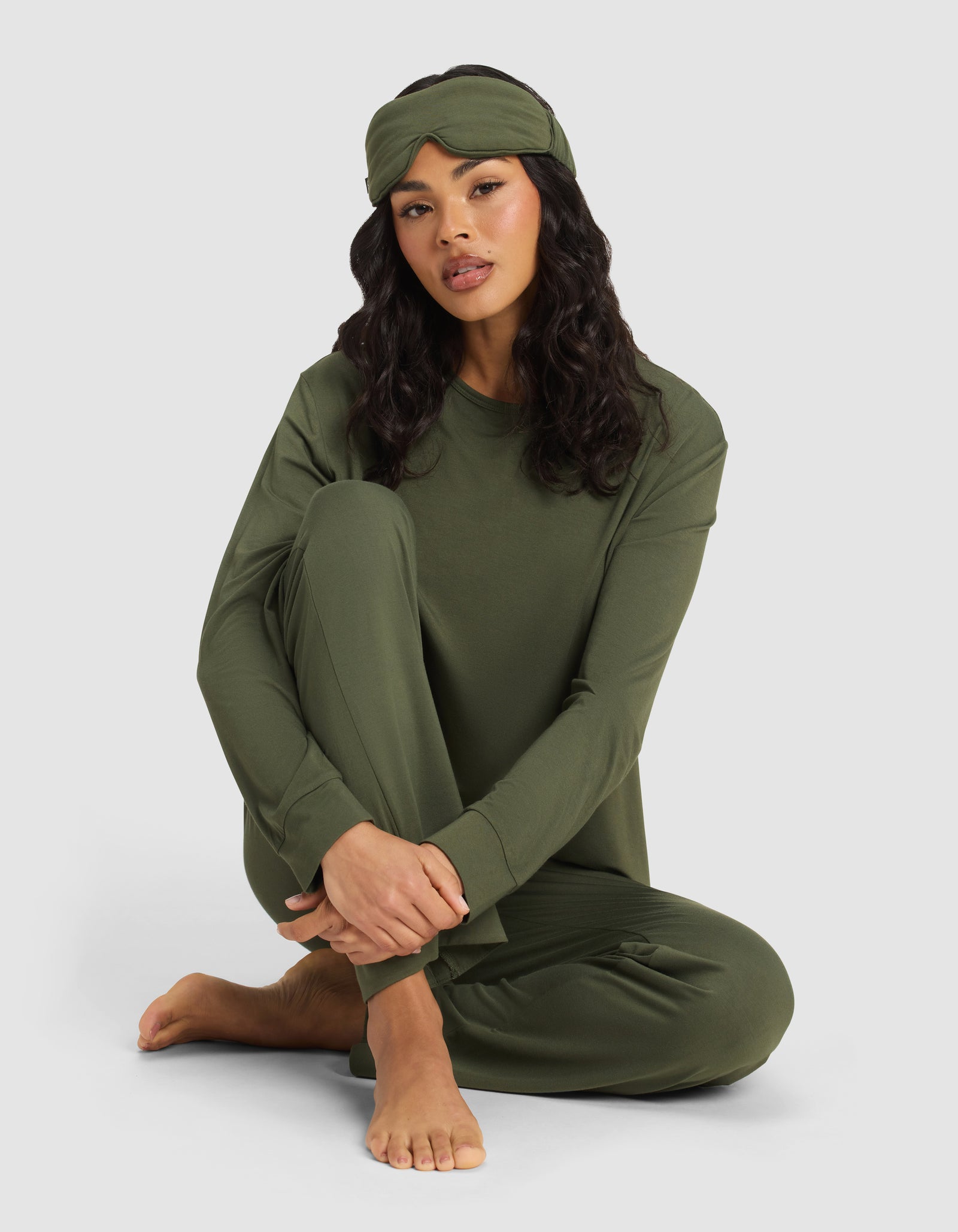 A woman with long dark hair sits barefoot on the floor, relaxed against a plain light background, wearing an olive green pajama set and the Cozy Earth Bamboo Stretch-Knit Sleep Mask on her head.