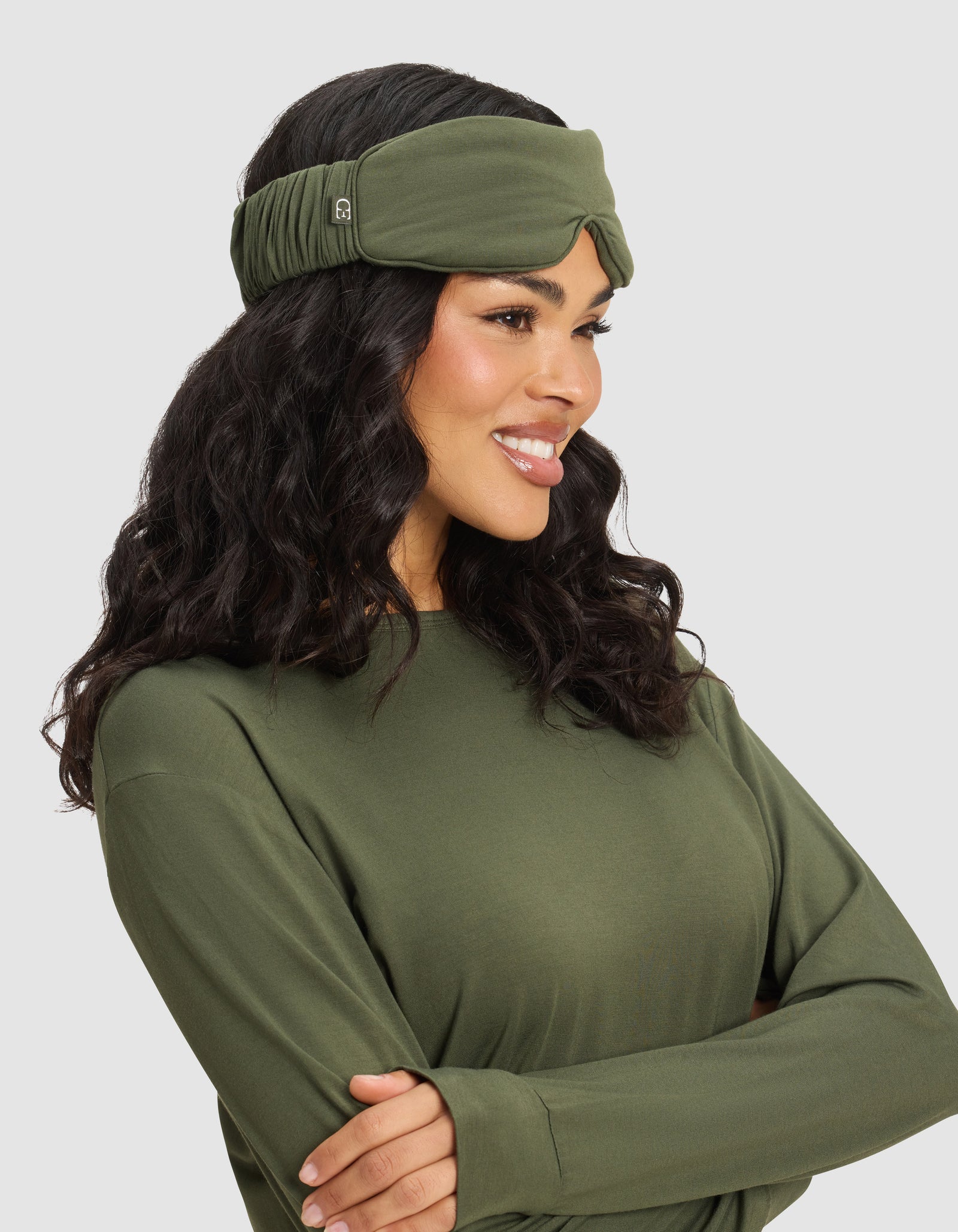 A woman with long, wavy dark hair smiles and looks to the side with arms folded, wearing a Cozy Earth Bamboo Stretch-Knit Sleep Mask on her head. She is set against a light gray background. 