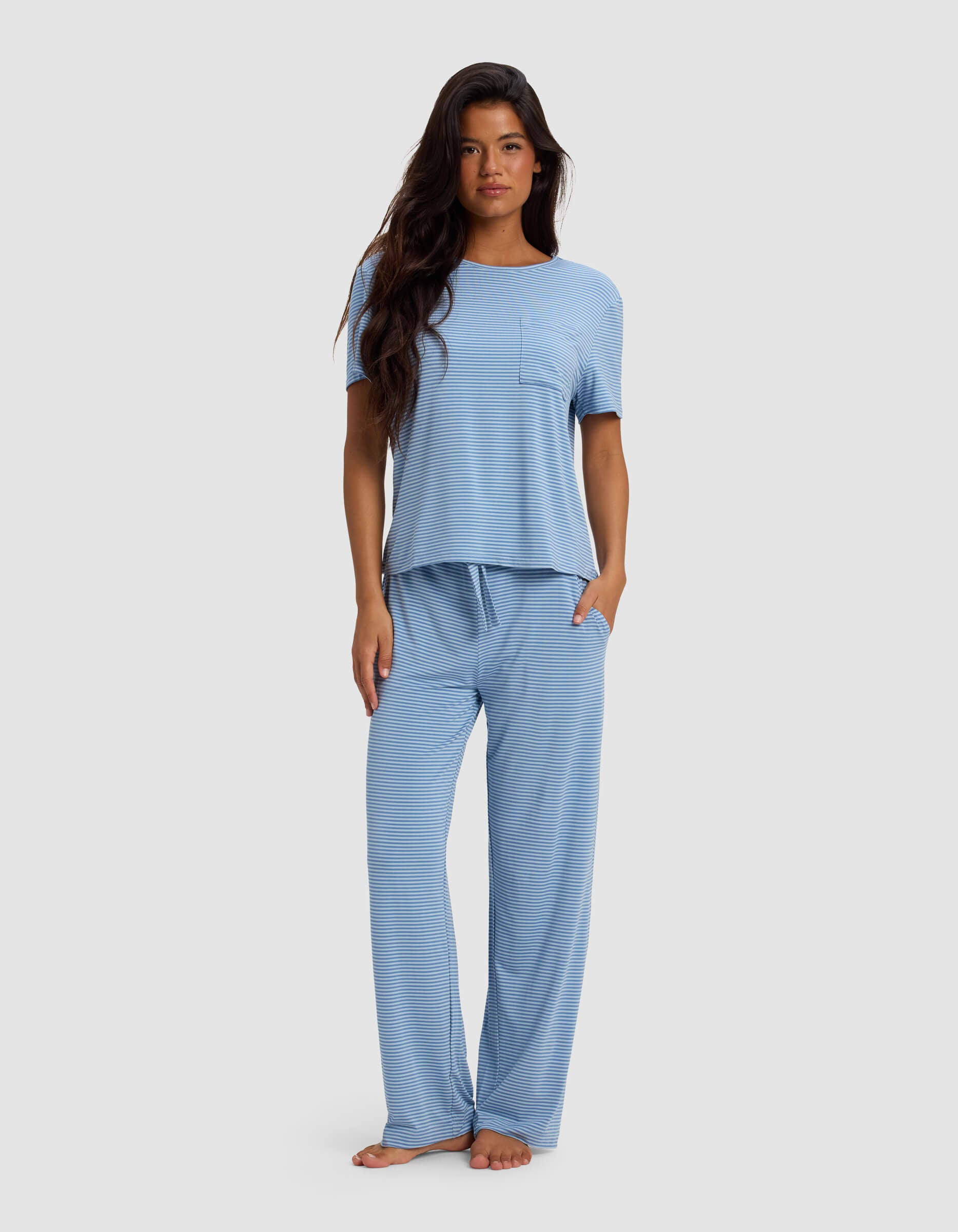 A woman with long dark hair stands barefoot against a plain backdrop, wearing the Cozy Earth Women's Bamboo Stretch-Knit Lounge Tee & Pant Set—a light blue, short-sleeve, striped pajama set featuring a chest pocket and loose-fit pants.