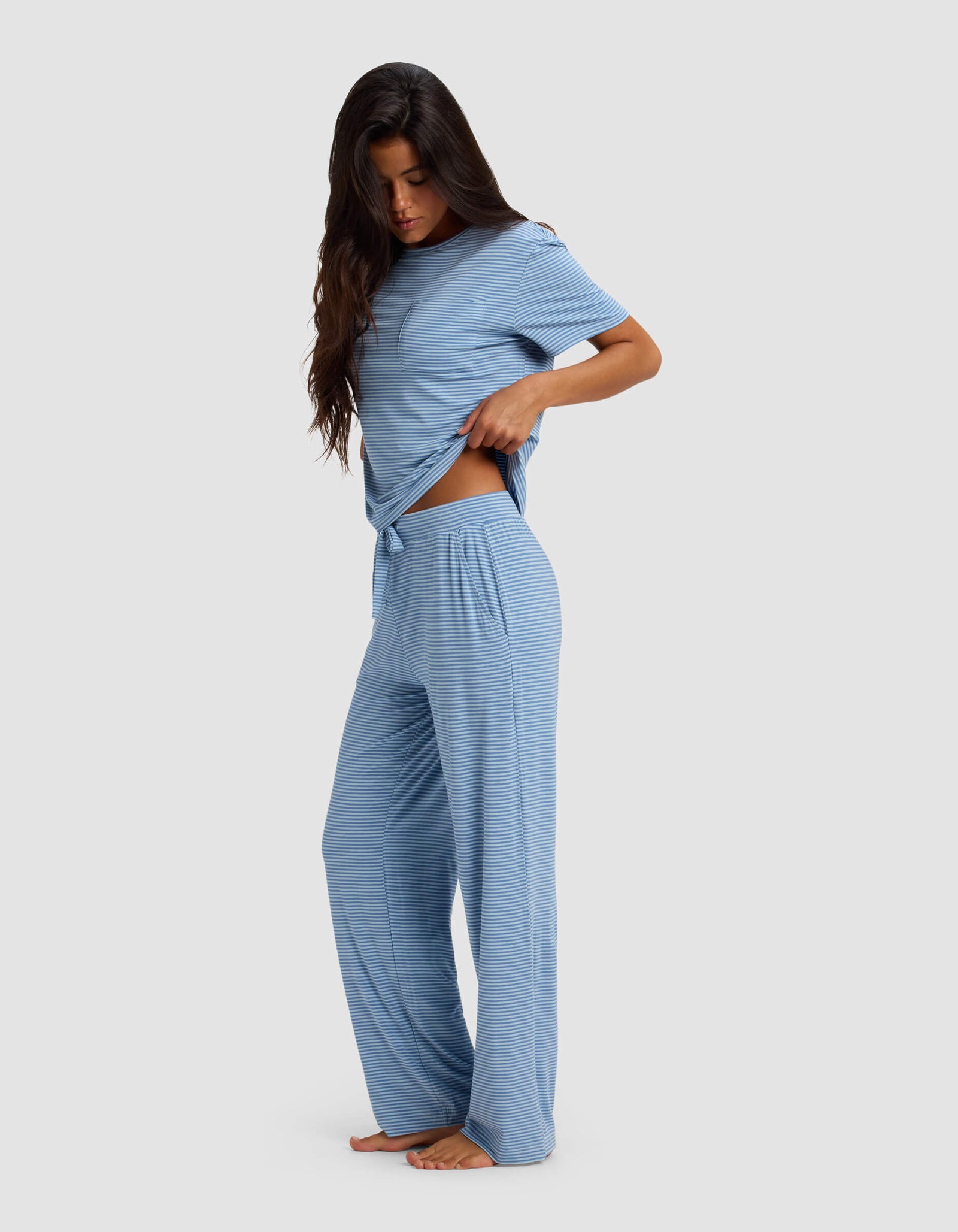 A woman with long dark hair wears the Cozy Earth Women's Bamboo Stretch-Knit Lounge Tee & Pant Set in light blue with subtle stripes. She stands barefoot, slightly lifting her top to reveal the matching pants against a light gray background.