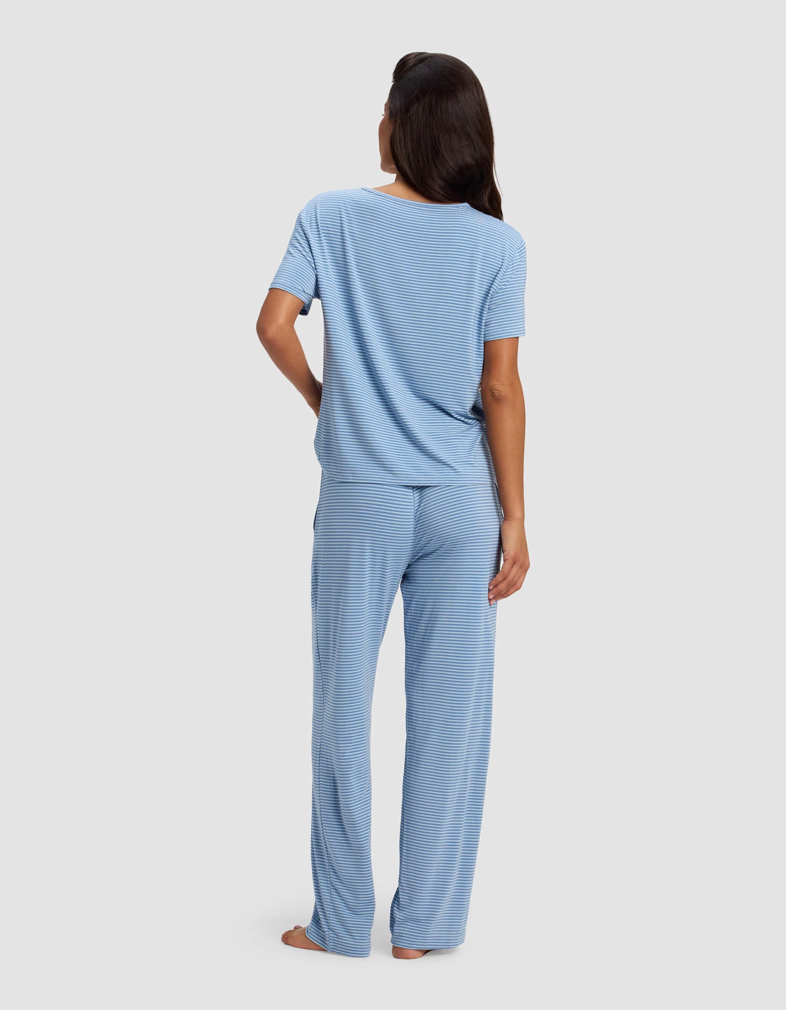 A woman stands barefoot facing away, wearing Cozy Earth's Women's Bamboo Stretch-Knit Lounge Tee & Pant Set in light blue with subtle horizontal stripes, against a plain light gray background.