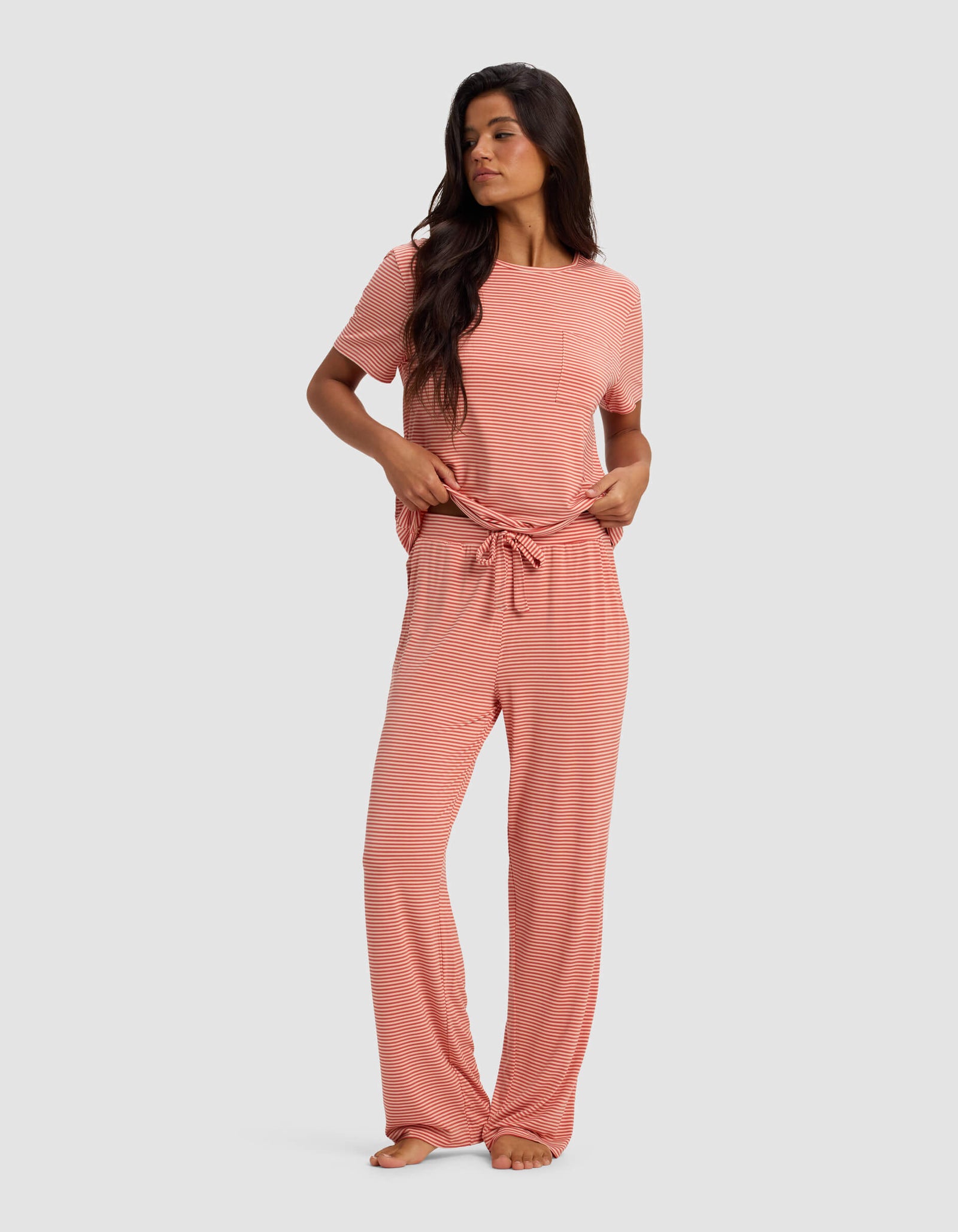 A woman in the Cozy Earth Women's Bamboo Stretch-Knit Lounge Tee & Pant Set, featuring pink and white stripes, short sleeves, front pocket, and drawstring waist, stands barefoot against a plain background, looking to the side.
