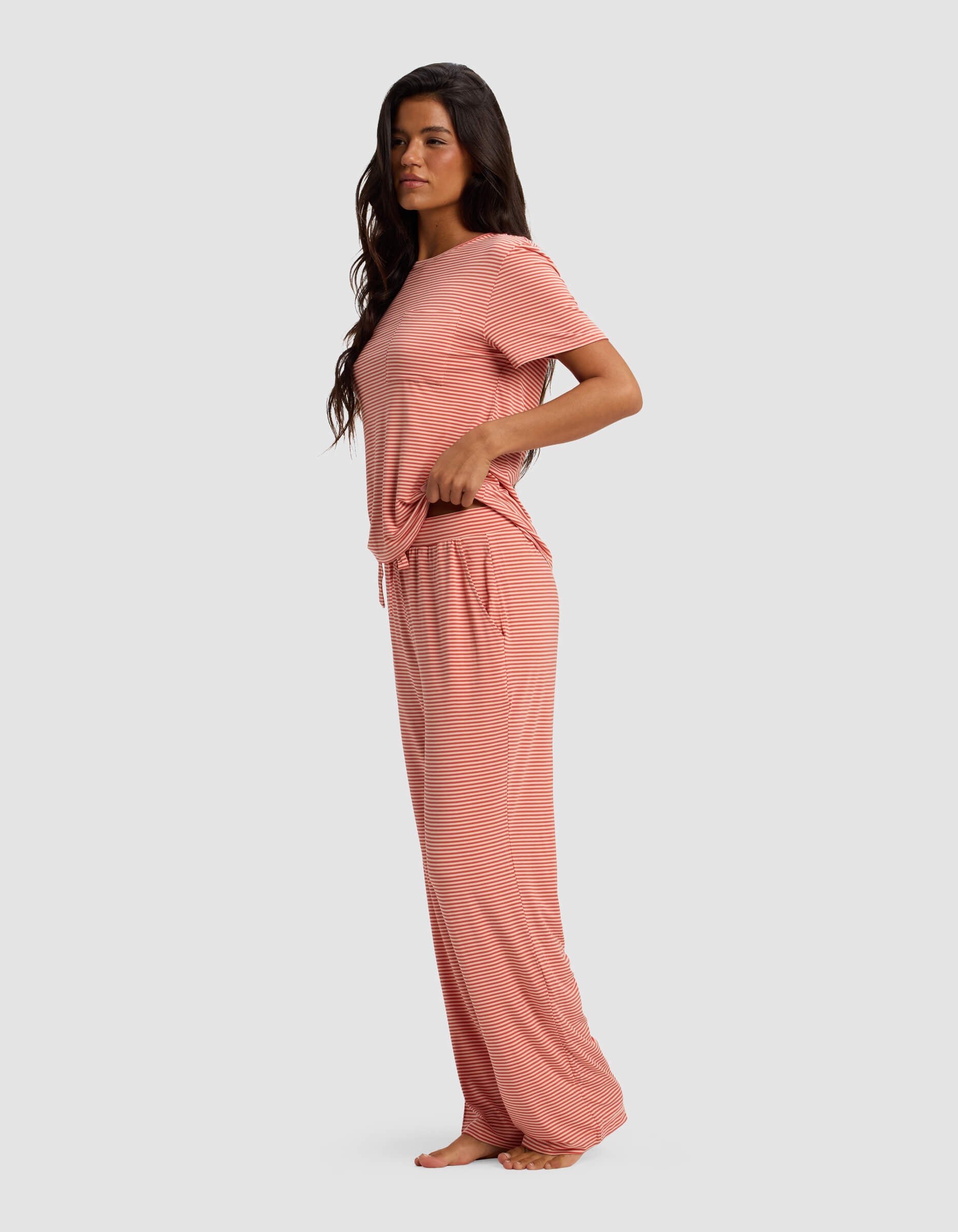 A woman stands barefoot in side profile, wearing Cozy Earth's Women's Bamboo Stretch-Knit Lounge Tee & Pant Set—loose-fitting with short sleeves and wide-leg pants—against a plain light gray background.