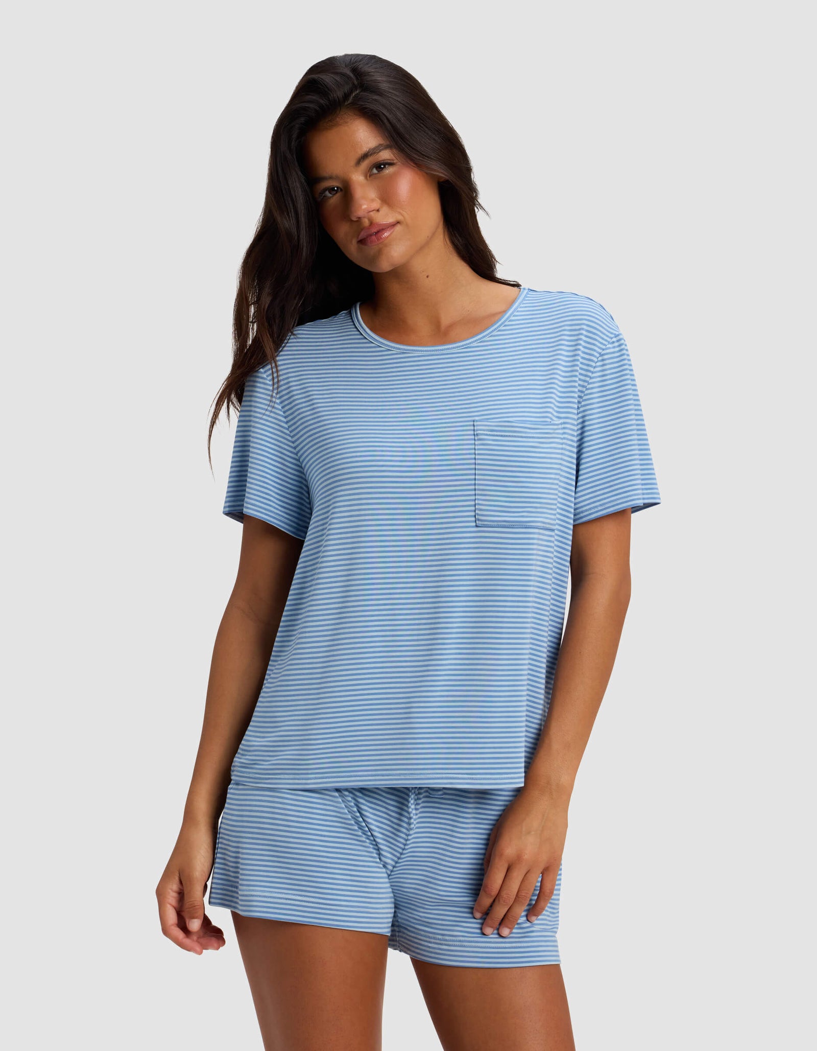 A woman wearing the Cozy Earth Women's Bamboo Stretch-Knit Lounge Tee in light blue and white stripes with a chest pocket and matching drawstring shorts stands against a plain light gray background.
