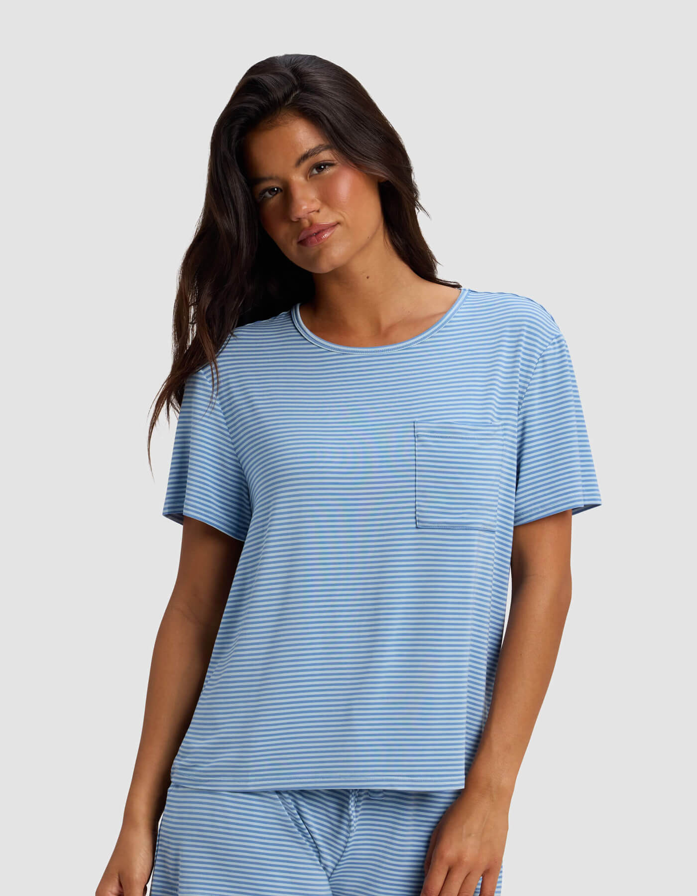A woman with long dark hair wears the Cozy Earth Women's Bamboo Stretch-Knit Lounge Tee & Pant Set, featuring a light blue and white striped short-sleeve top with a chest pocket, standing against a plain light gray background.