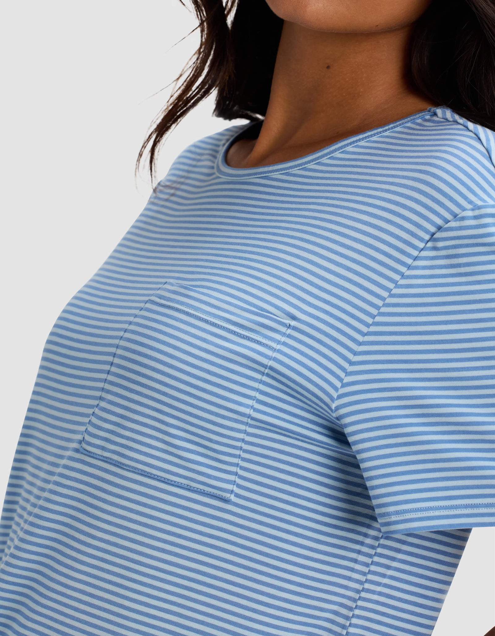 A woman wears the Cozy Earth Women's Bamboo Stretch-Knit Lounge Tee in light blue and white stripes with a chest pocket. The image is cropped to highlight the shirt's design and texture on her upper body.