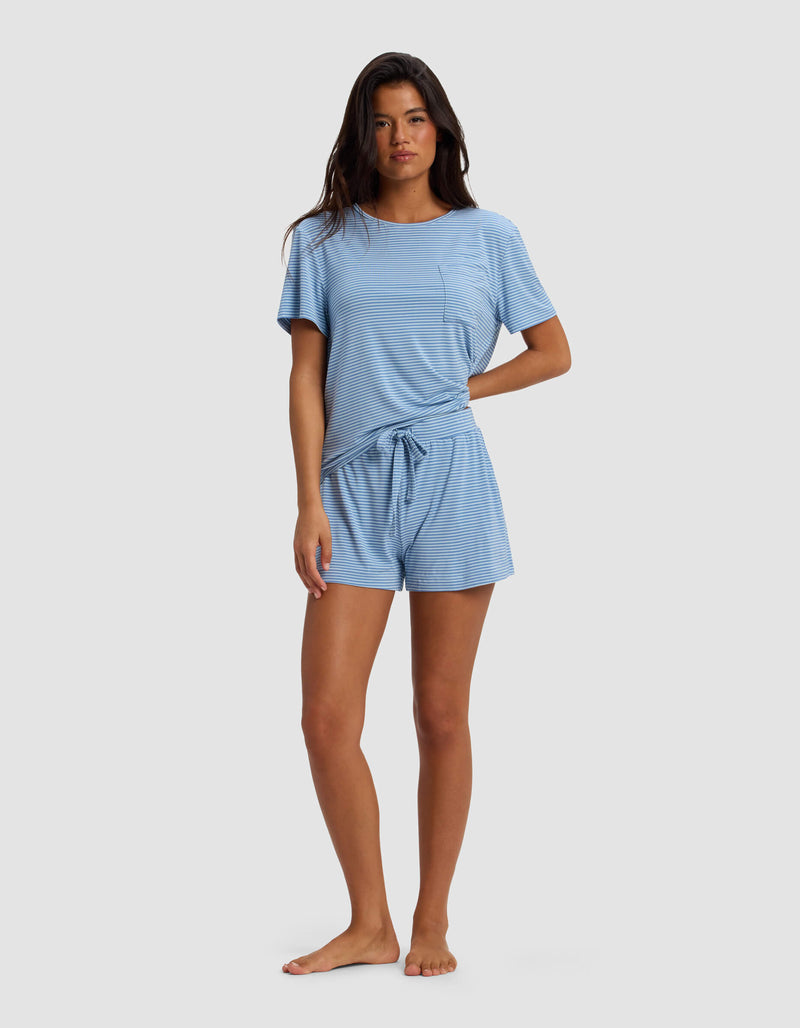 A woman stands barefoot against a plain background, wearing the Cozy Earth Women's Bamboo Stretch-Knit Lounge Tee & Short Set in light blue with short sleeves, subtle stripes, a chest pocket, and a waist tie.