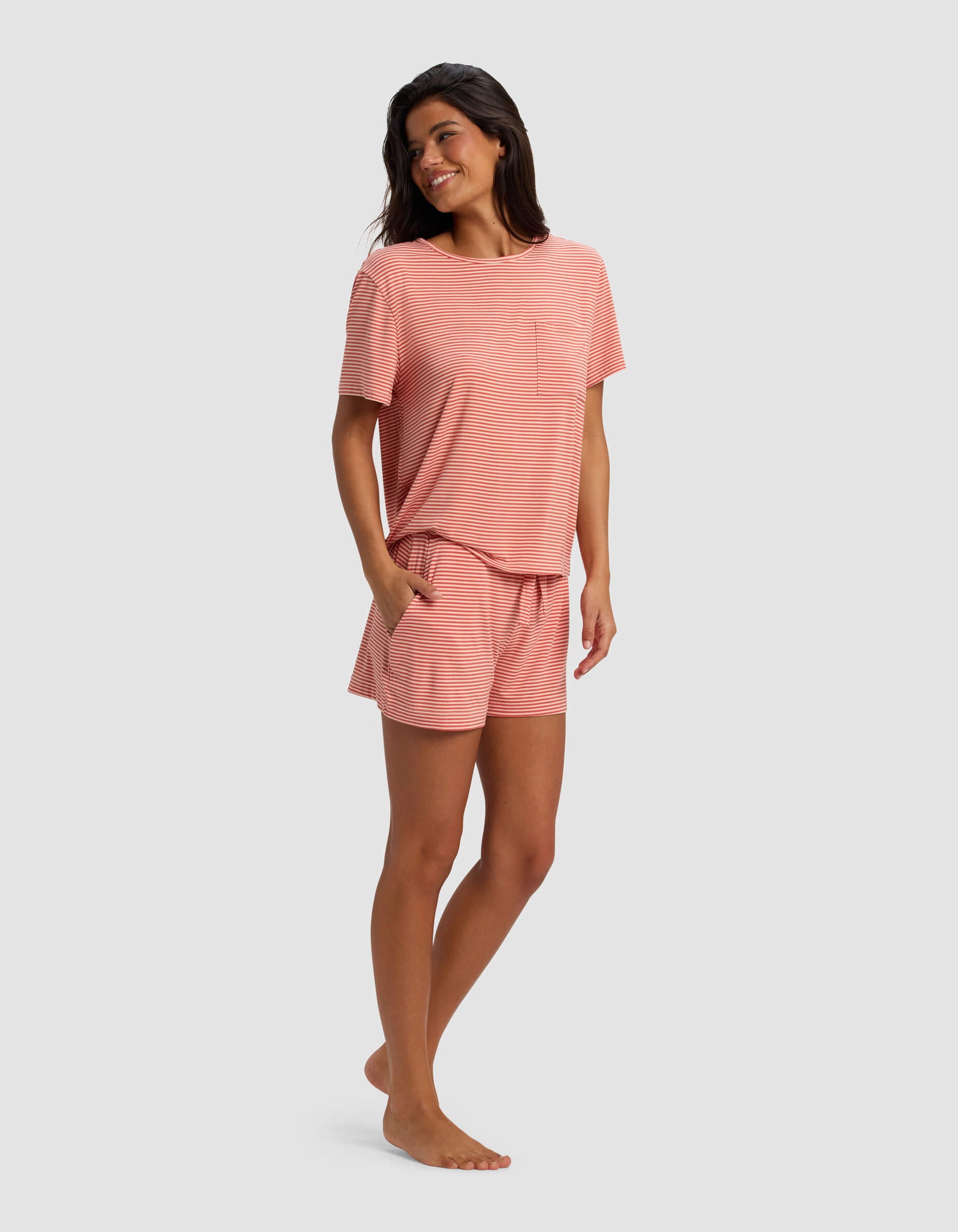 A woman stands barefoot, smiling in the Cozy Earth Women's Bamboo Stretch-Knit Lounge Tee & Short Set—loose-fitting, short-sleeved, coral pink striped top and matching shorts with pockets—against a plain light background.