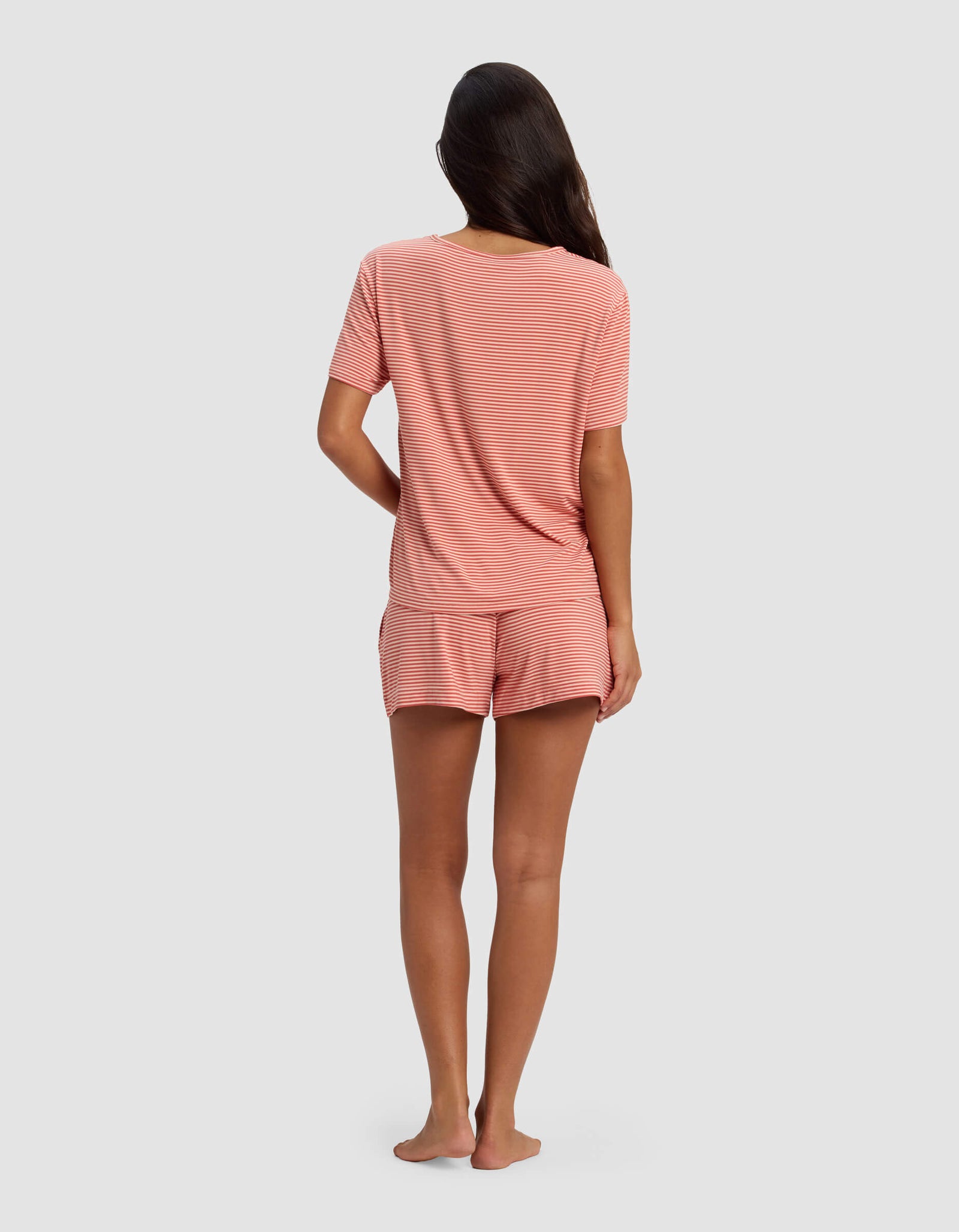 A woman with long dark hair stands barefoot, facing away, wearing the Cozy Earth Women's Bamboo Stretch-Knit Lounge Tee & Short Set in pink and white stripes, against a plain light gray background.