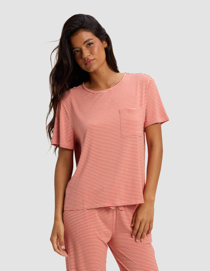 A woman with long dark hair wears the Cozy Earth Women's Bamboo Stretch-Knit Lounge Tee in coral and white stripes, featuring a small chest pocket, while standing against a plain light gray background.