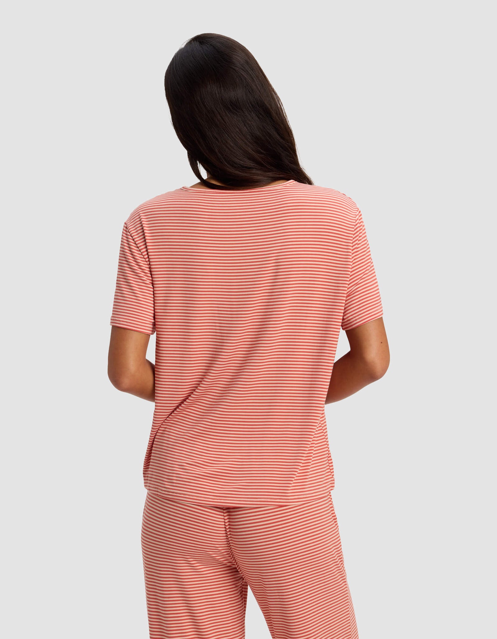 A woman with long dark hair, seen from behind, models the Cozy Earth Women's Bamboo Stretch-Knit Lounge Tee in a short-sleeved red and white striped style, paired with matching pants, against a plain light gray background.