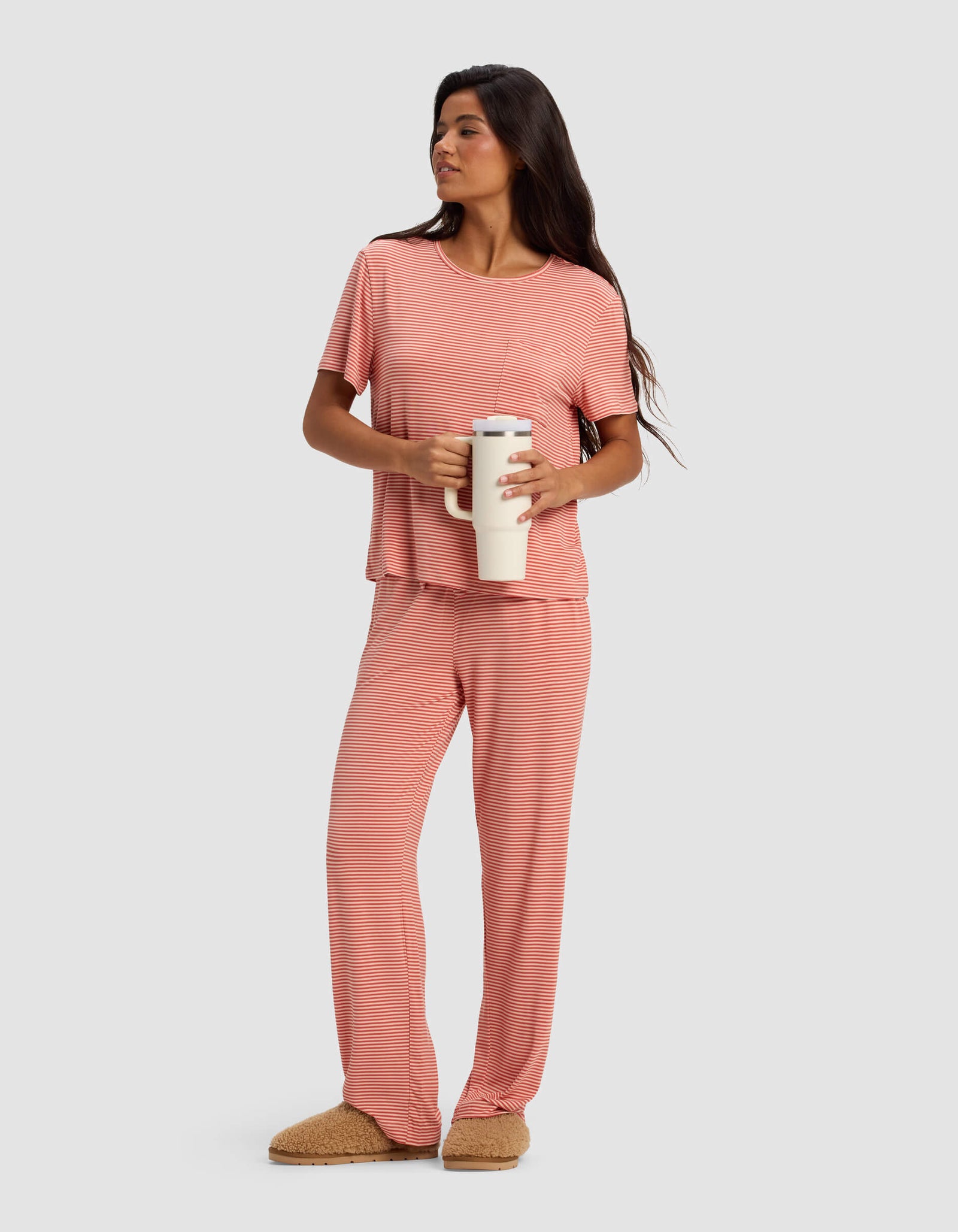 A woman with long dark hair wears the Cozy Earth Women's Bamboo Stretch-Knit Lounge Tee in pink stripes, paired with tan slippers and holding a white travel mug. She stands against a light gray background, looking to the side with a slight smile.