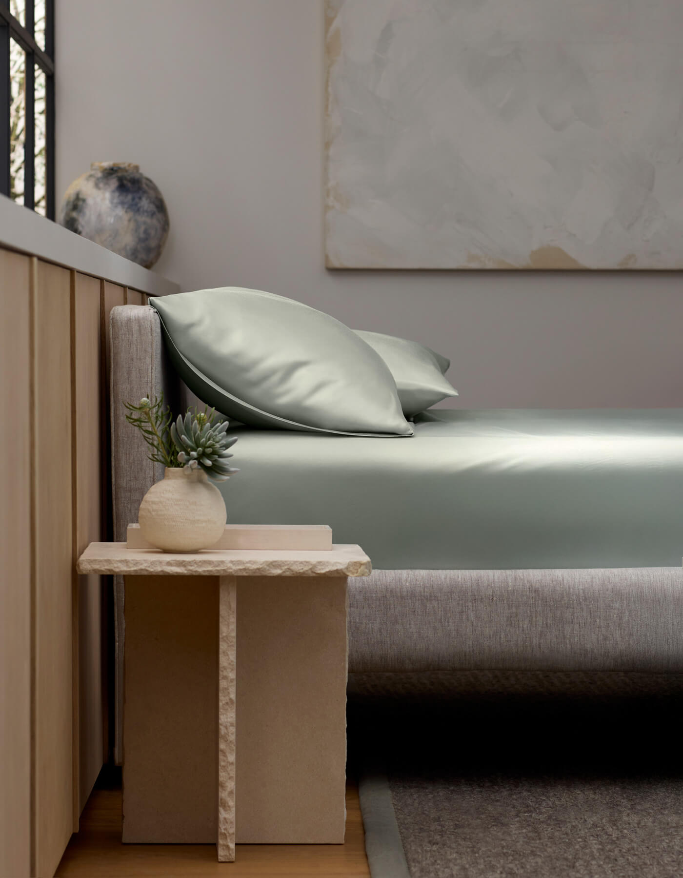 A minimalist bedroom with a light gray upholstered bed, Cozy Earth’s Bamboo Bedding Basic Bundle in sage green, and two pillows. A stone side table holds a potted succulent. Neutral wall art and a vase accent the softly lit background.