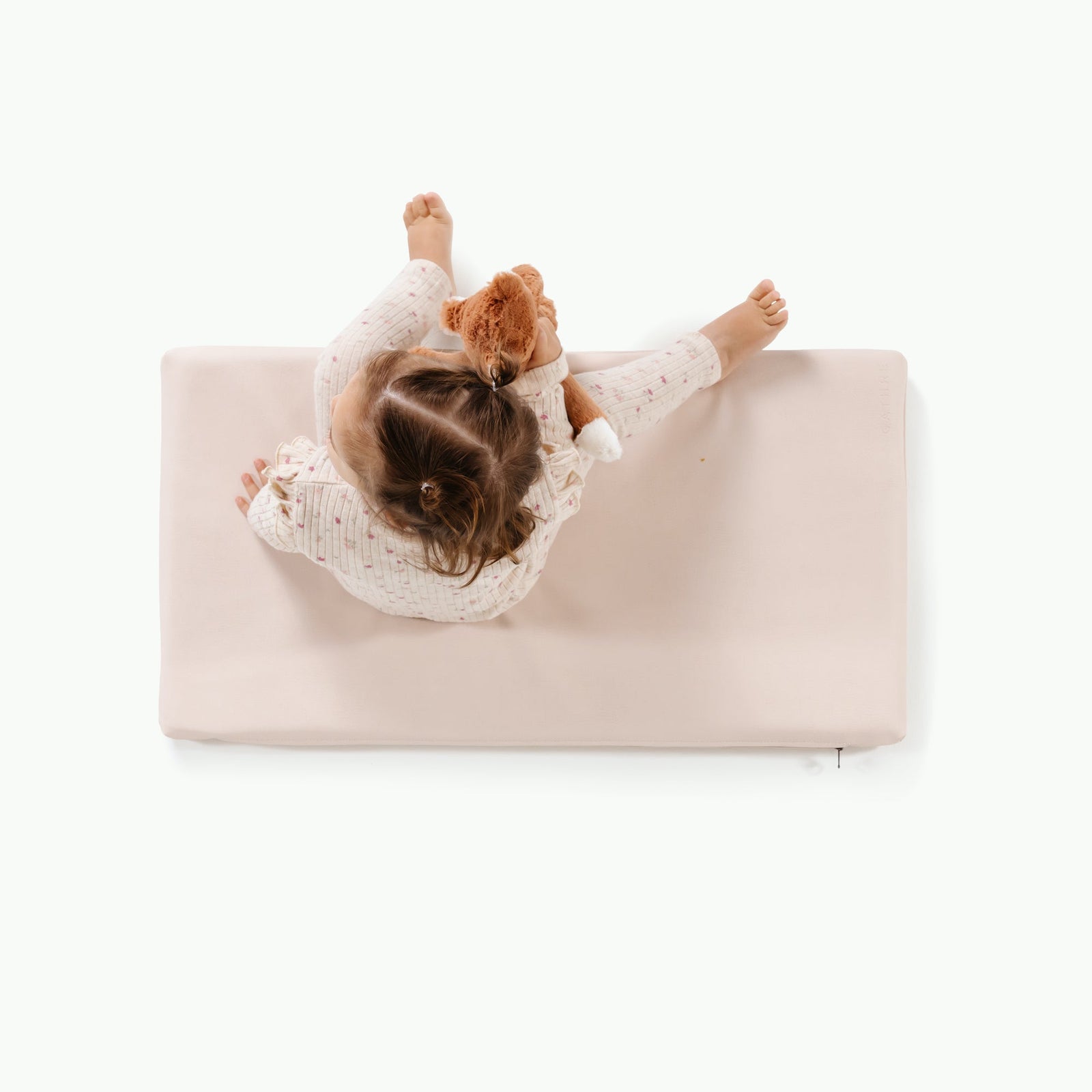 A toddler sits on the Gathre Wholesale Padded Changing Mat, wearing light pajamas and holding a brown stuffed animal, viewed from above against a white background.