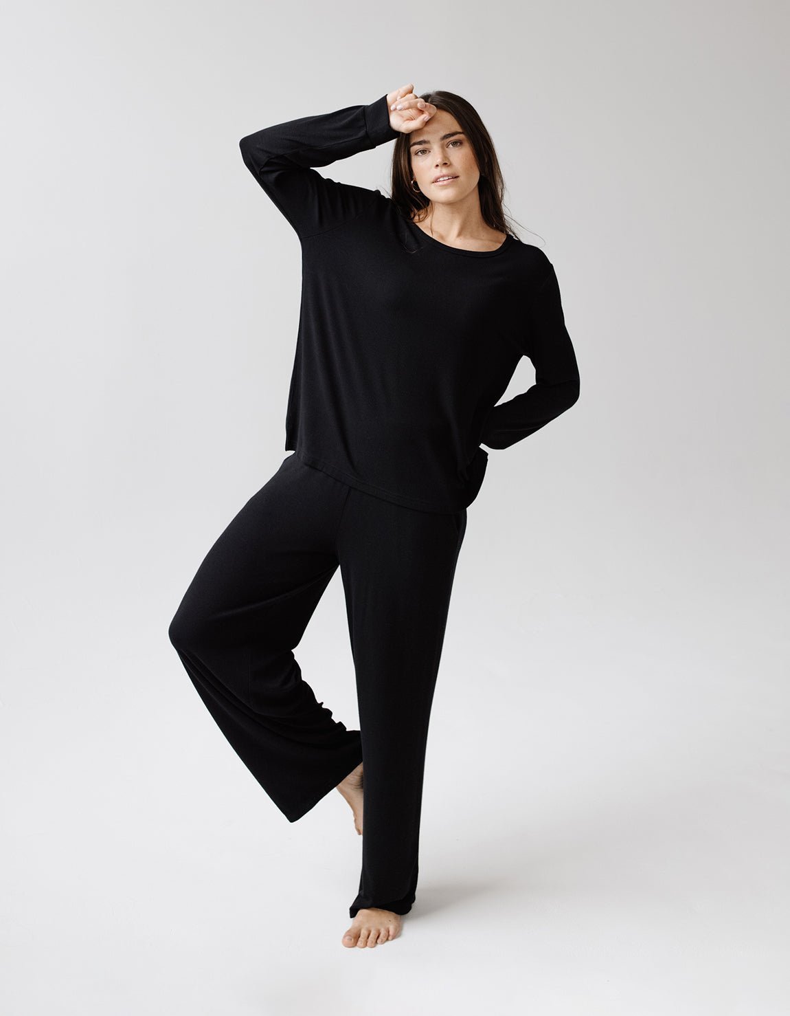 A woman stands barefoot on one leg in a relaxed pose against a plain light gray background, wearing Cozy Earth's Women's Bamboo Rib-Knit Lounge Pant and a long-sleeve black top.