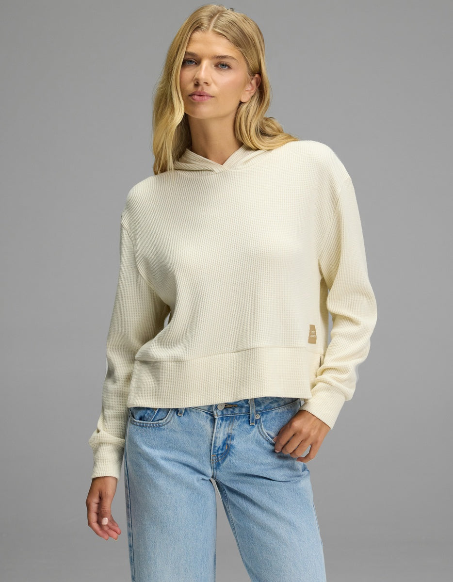 A person wearing a cream-colored Women’s Blake Waffle Hoodie with a small rectangular tag on the side, paired with light-wash blue jeans.