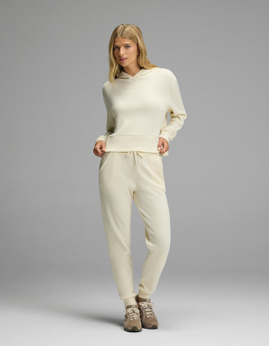 A woman wearing a cream-colored Women’s Blake Waffle Hoodie & Jogger Set featuring a hooded long-sleeve top and matching joggers with a textured knit pattern.|Color:Coconut