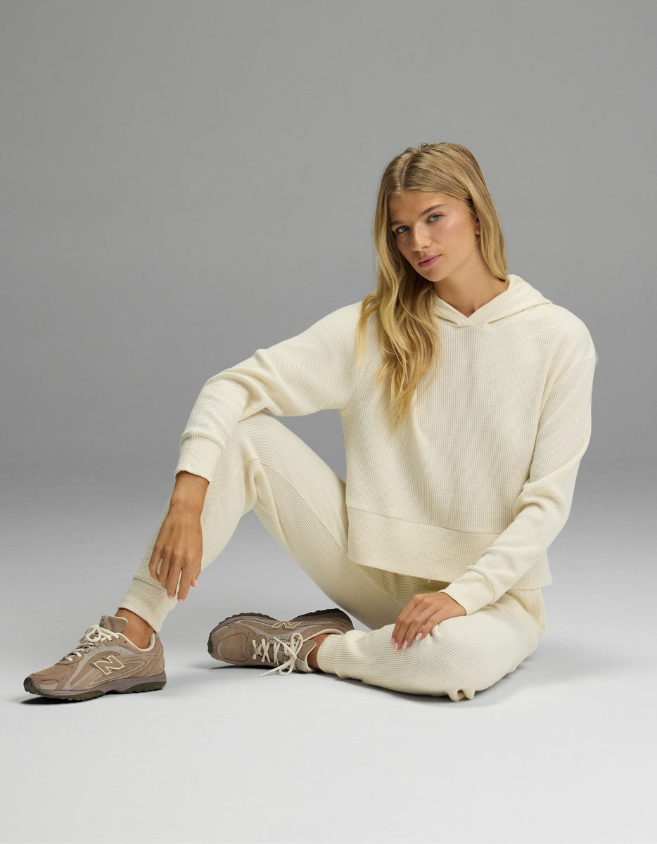 A woman sits on the floor wearing a cream-colored Women’s Blake Waffle Hoodie & Jogger Set with matching beige athletic sneakers.