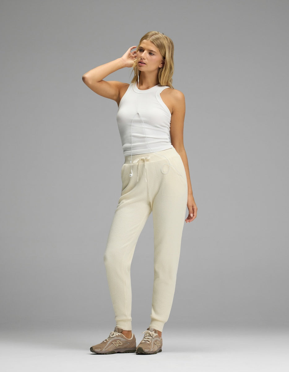 A woman wearing a white ribbed tank top and cream-colored Women’s Blake Waffle Jogger pants with a drawstring waist and cuffed ankles, paired with brown athletic sneakers.