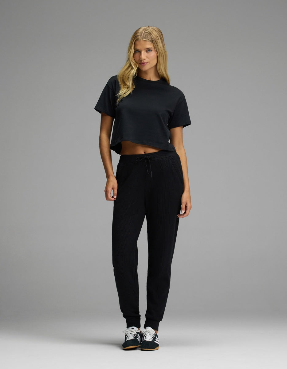 A woman wears a black cropped t-shirt and black Cozy Earth Women’s Blake Waffle Joggers with a drawstring waist and ribbed texture, paired with black and white sneakers.