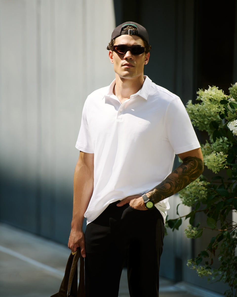 A man wearing a white Cozy Earth Everday Polo shirt, black pants, a black cap, and sunglasses, standing outdoors next to a gray wall and green foliage. 
