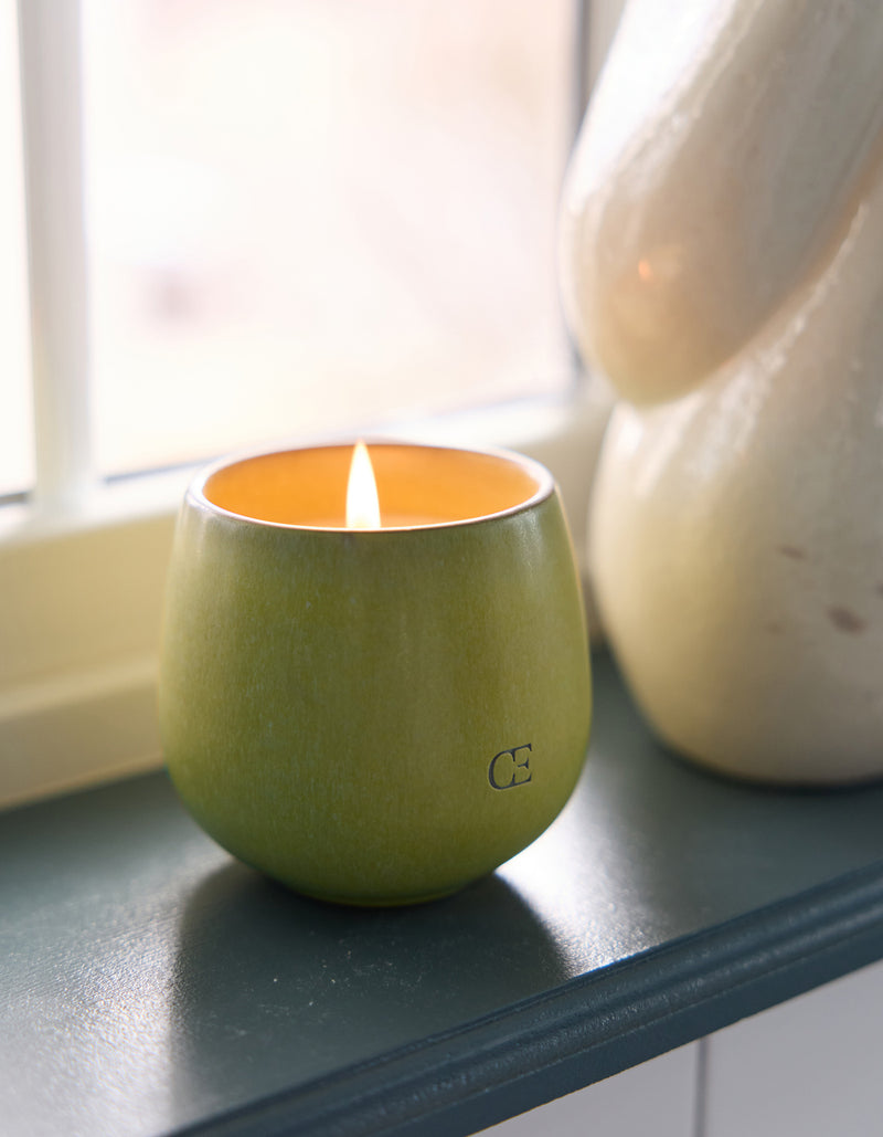 A Candle BOTANIC by Cozy Earth in a round holder burns on a windowsill, accompanied by a light-yellow ceramic piece. Natural light streams through the window in the background.