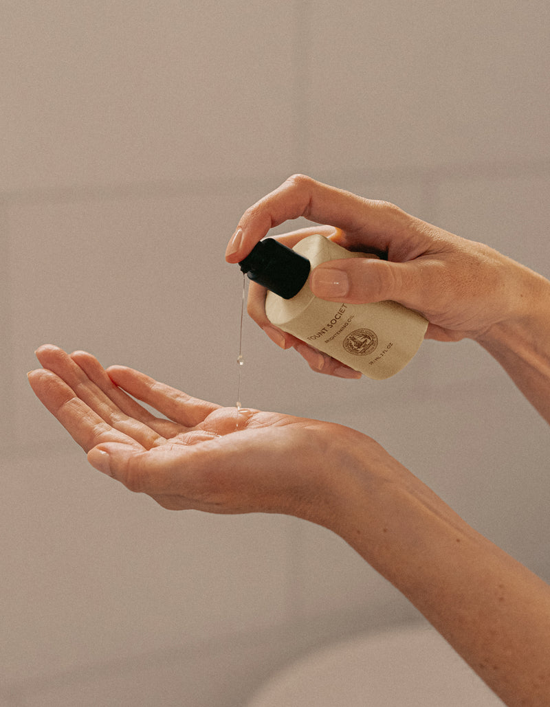 A person dispenses Cozy Earth's Brightening Vitamin C Oil from a small bottle with a black pump onto their open palm, emphasized against a softly blurred background.