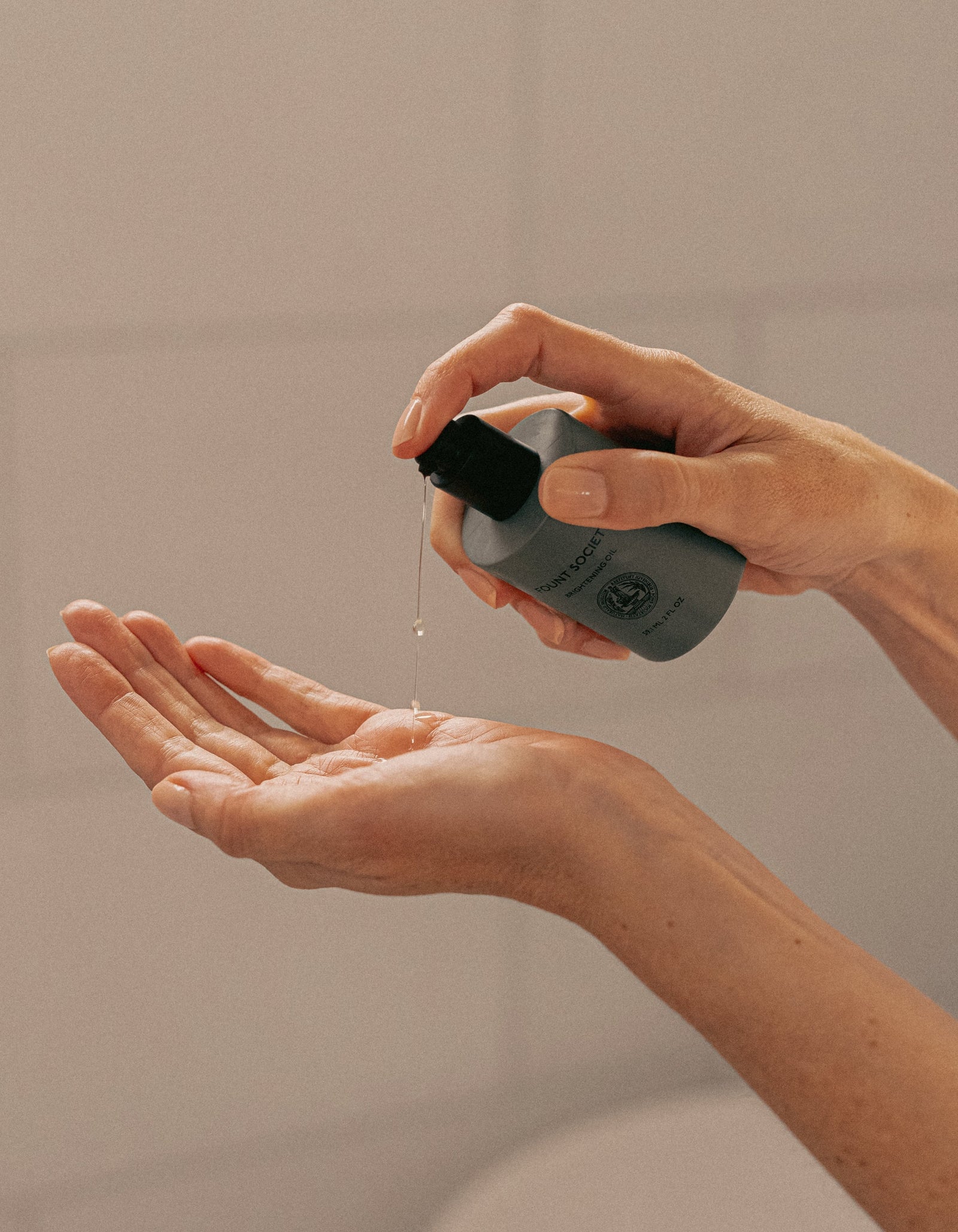 A person squeezes liquid from a small, gray bottle labeled Day-to-Night Essentials by Cozy Earth onto their open palm, with hands set against a softly blurred background.