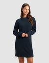 A woman with long brown hair wears the Cozy Earth Women's Brooklyn Sweater Dress in dark blue. She stands against a light gray background, looking to her left with a slight smile and her hands together in front of her.|Color:Eclipse