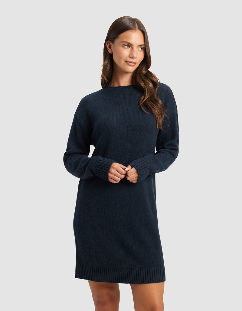 A woman with long brown hair wears the Cozy Earth Women's Brooklyn Sweater Dress in dark blue. She stands against a light gray background, looking to her left with a slight smile and her hands together in front of her.