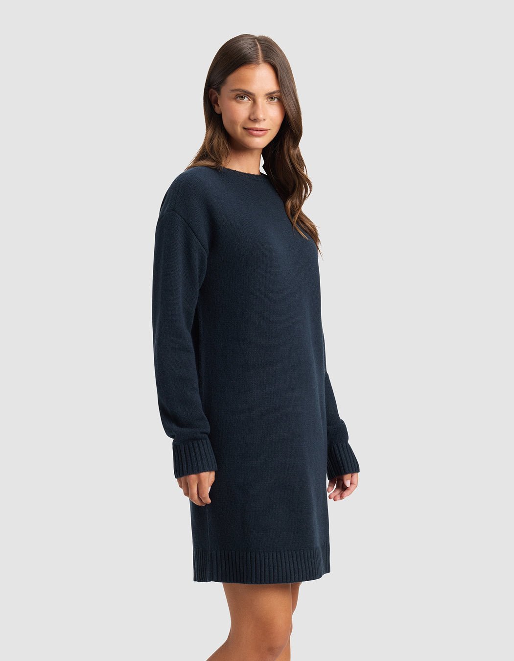 A woman with long brown hair models the Cozy Earth Women's Brooklyn Sweater Dress, a loose-fitting, long-sleeve navy blue piece, standing against a plain light gray background and smiling gently.|Color:Eclipse