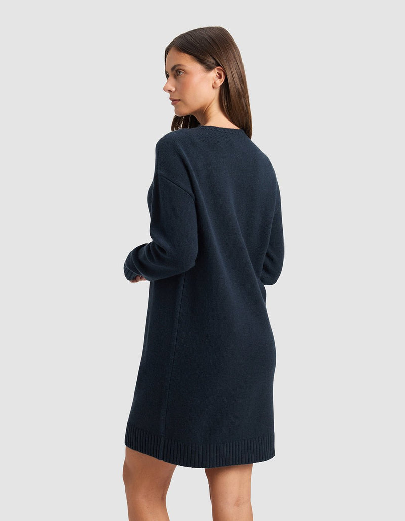 A woman with straight brown hair models the Cozy Earth Women's Brooklyn Sweater Dress in dark navy, featuring long sleeves. She stands sideways, looking left, against a plain light gray background.