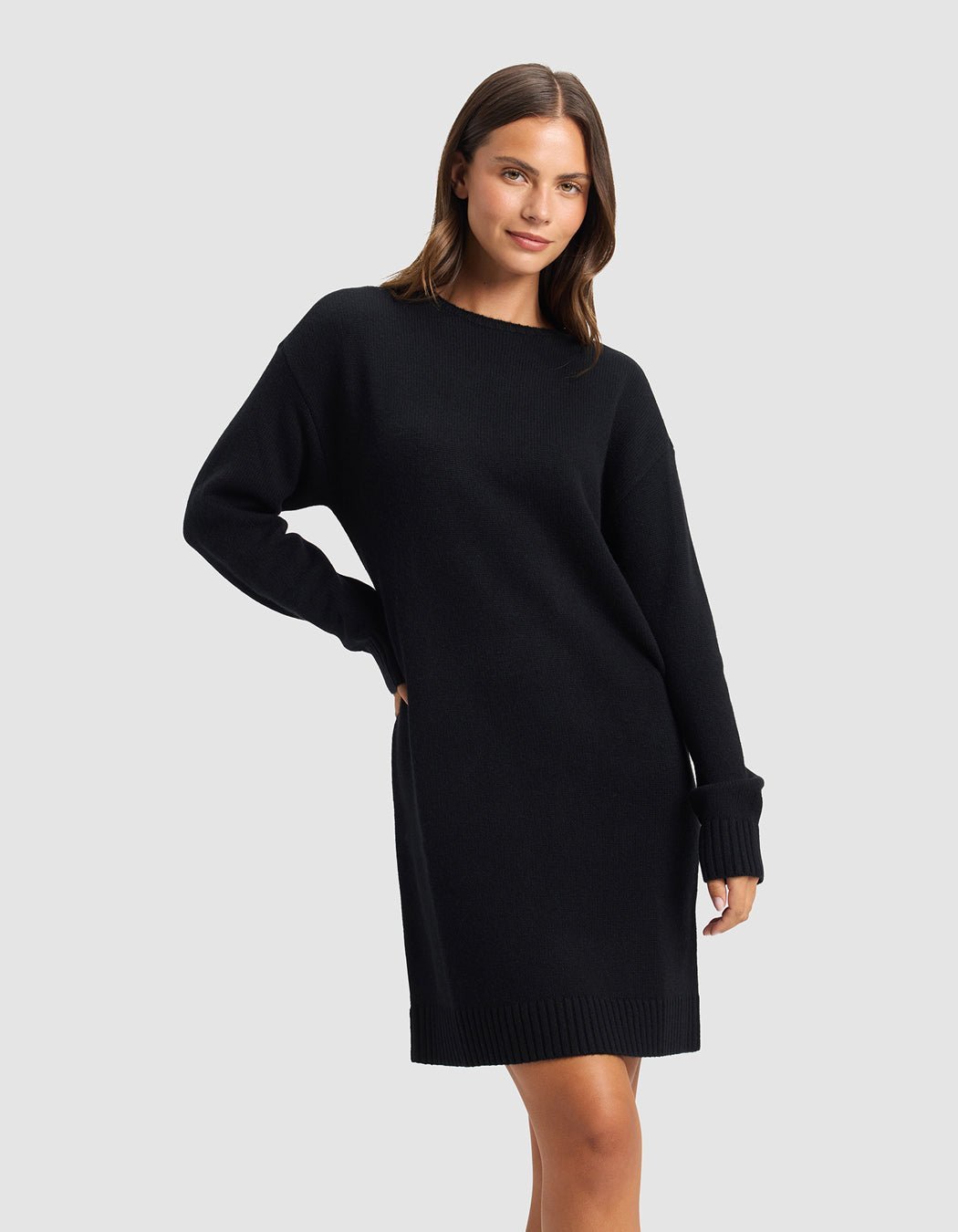 A woman poses against a light gray background, wearing the Cozy Earth Women's Brooklyn Sweater Dress—a loose-fitting, long-sleeved black knit style. She smiles softly with one hand on her hip.|Color:Jet Black