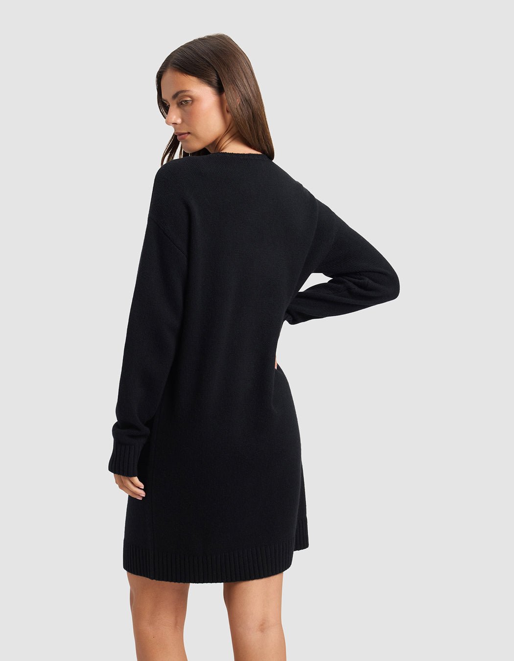A woman wearing the Cozy Earth Women's Brooklyn Sweater Dress in black stands with her left hand on her hip, looking over her shoulder against a plain light gray background.|Color:Jet Black