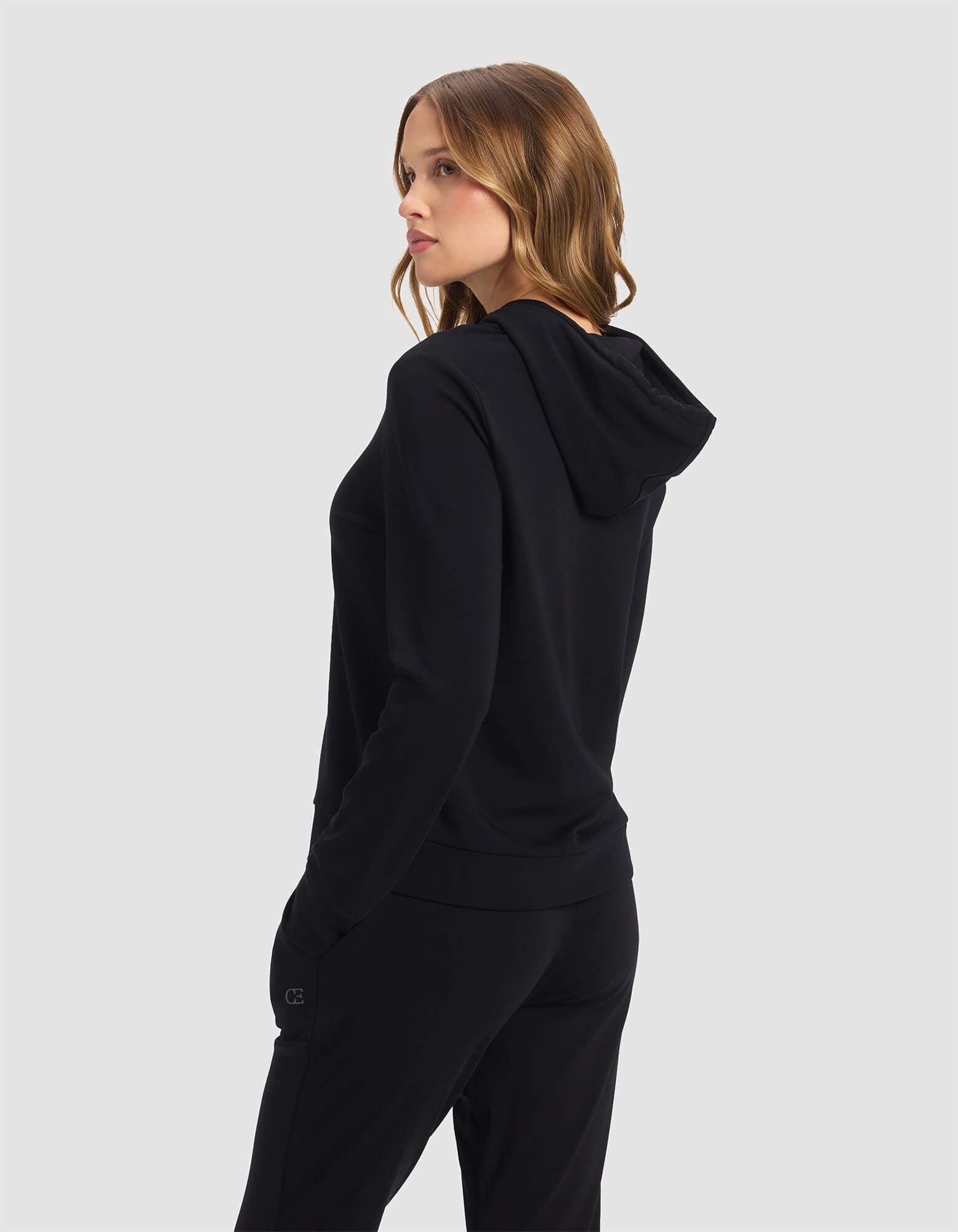 A woman with wavy, light brown hair models the Cozy Earth Women's Brushed Bamboo Hoodie in black, standing sideways and looking over her shoulder against a plain gray background.