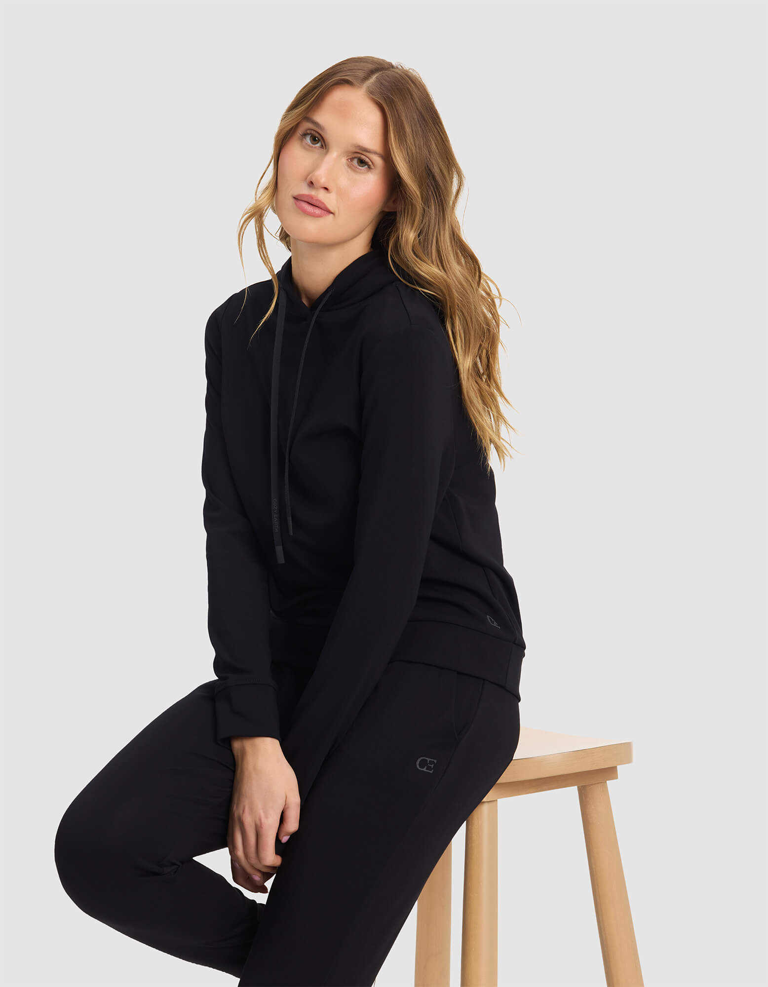 A woman with long, wavy blonde hair sits on a wooden stool against a plain light gray background, wearing the Cozy Earth Women's Brushed Bamboo Hoodie and black pants. She looks at the camera with a neutral expression.