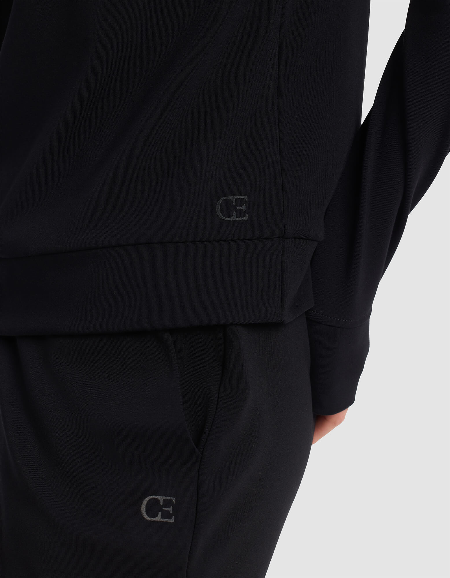 A person wears the Cozy Earth Women's Brushed Bamboo Hoodie in black, featuring subtle "CE" logos near the hem; their arm rests by their side, showing only part of their torso and legs.